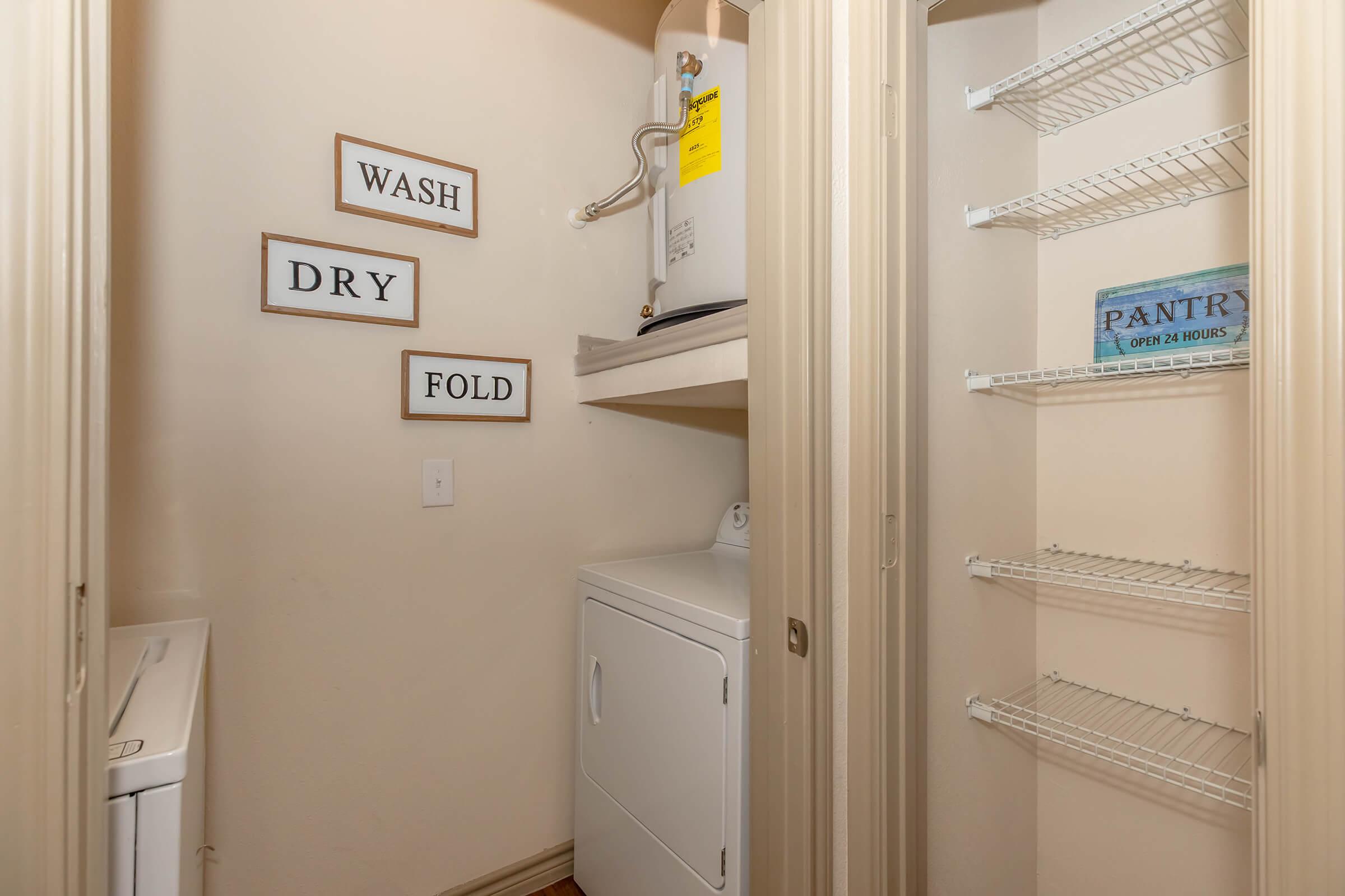A small laundry room featuring a washing machine and dryer. The walls have decorative signs that read "WASH," "DRY," and "FOLD." There is a water heater on a shelf, and a closet with wire shelves labeled "PANTRY." The room has a light-colored interior and wooden flooring.