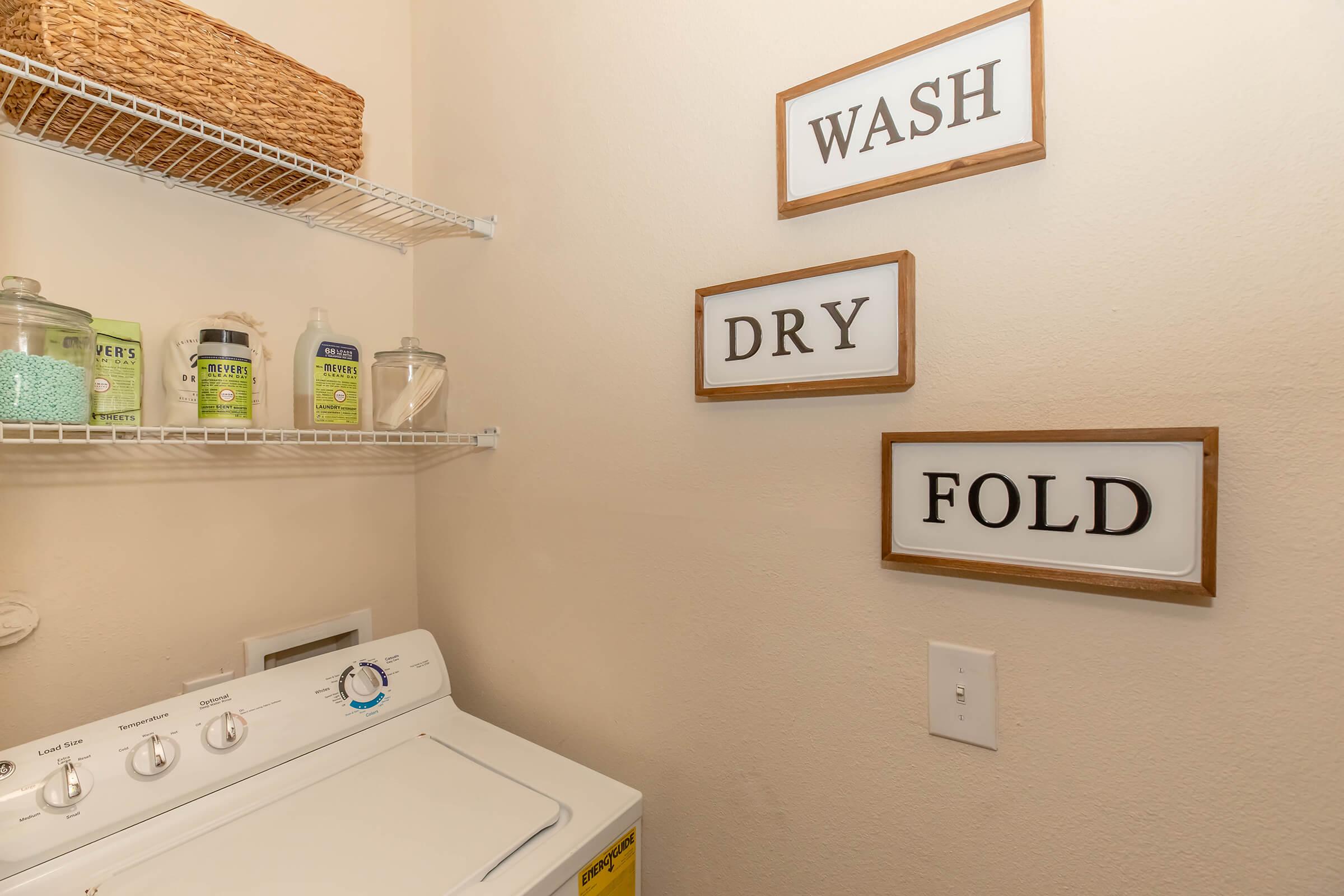 A laundry room featuring a washing machine, with decorative signs on the wall that read "WASH," "DRY," and "FOLD." There’s also a wire shelf above the machine holding baskets and various cleaning supplies. The walls are light-colored and the overall space is tidy and organized.