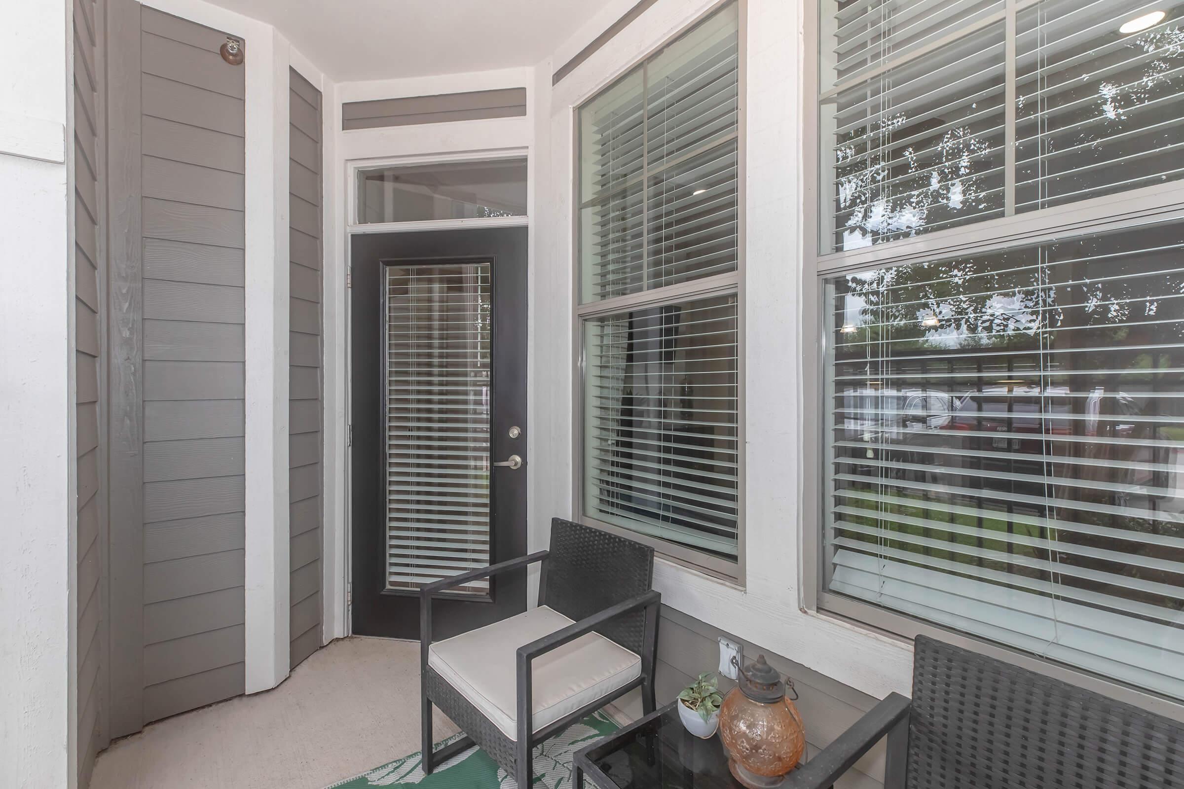 A cozy entryway featuring a small seating area with two chairs and a glass table. The space includes large windows with blinds, allowing natural light to fill the area. The walls are a mix of light and dark gray siding, creating a modern look. A potted plant and decorative items add a touch of personality.