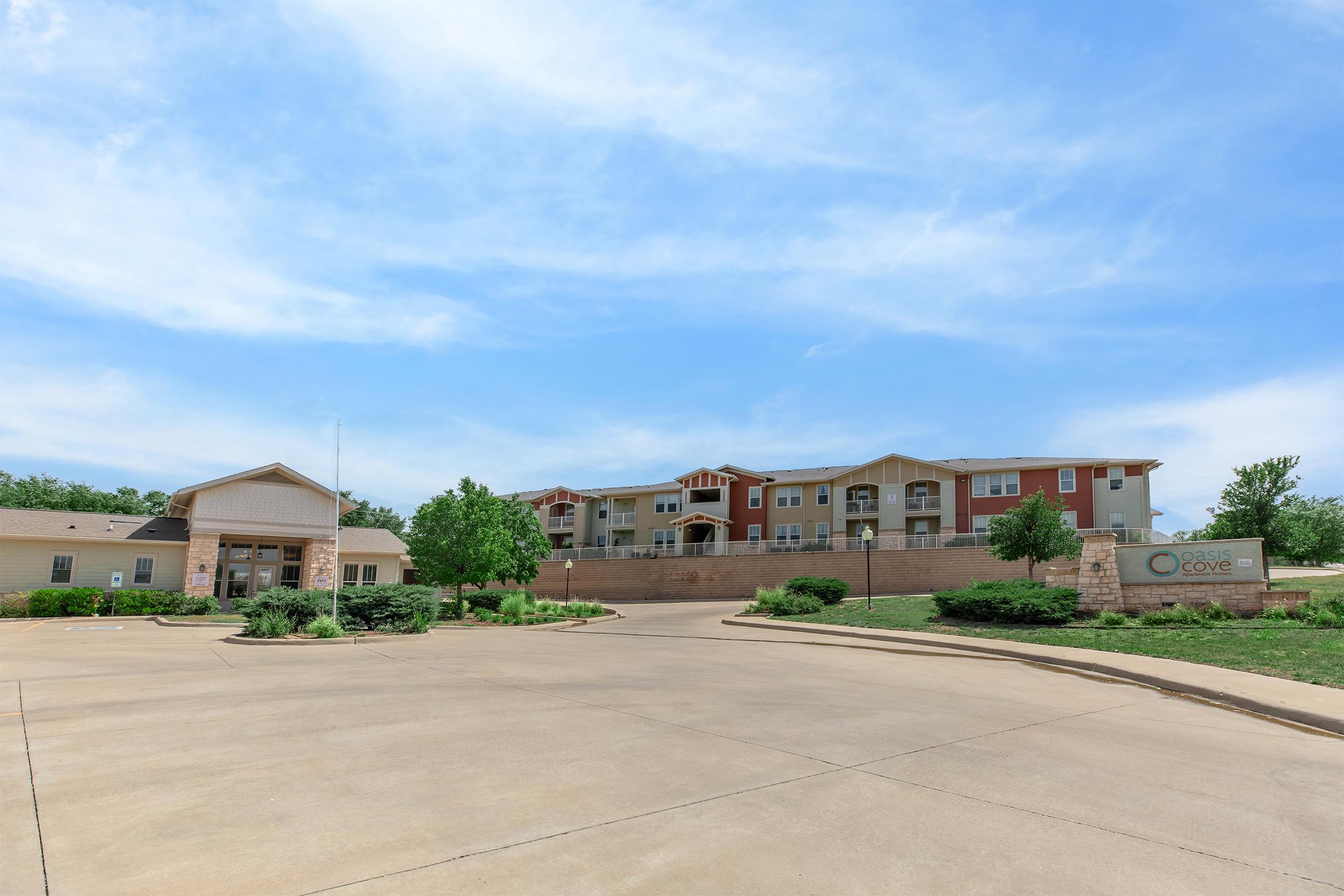 A residential apartment complex with a modern design, featuring multiple buildings. The main office structure is in the foreground with a landscaped entrance. The background includes larger apartment buildings. The sky is partly cloudy, and the area is well-maintained with greenery.