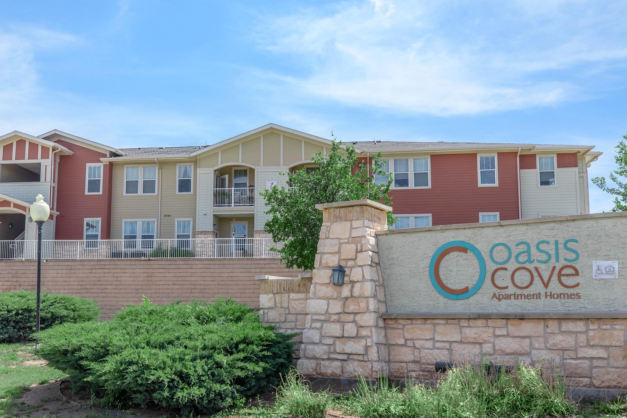 A well-maintained apartment complex named "Oasis Cove Apartment Homes." The building features a mix of beige, yellow, and red siding, with multiple balconies. A landscaped area with shrubs is in the foreground, and a stone sign displaying the complex's name is prominently featured. The sky is clear and blue.