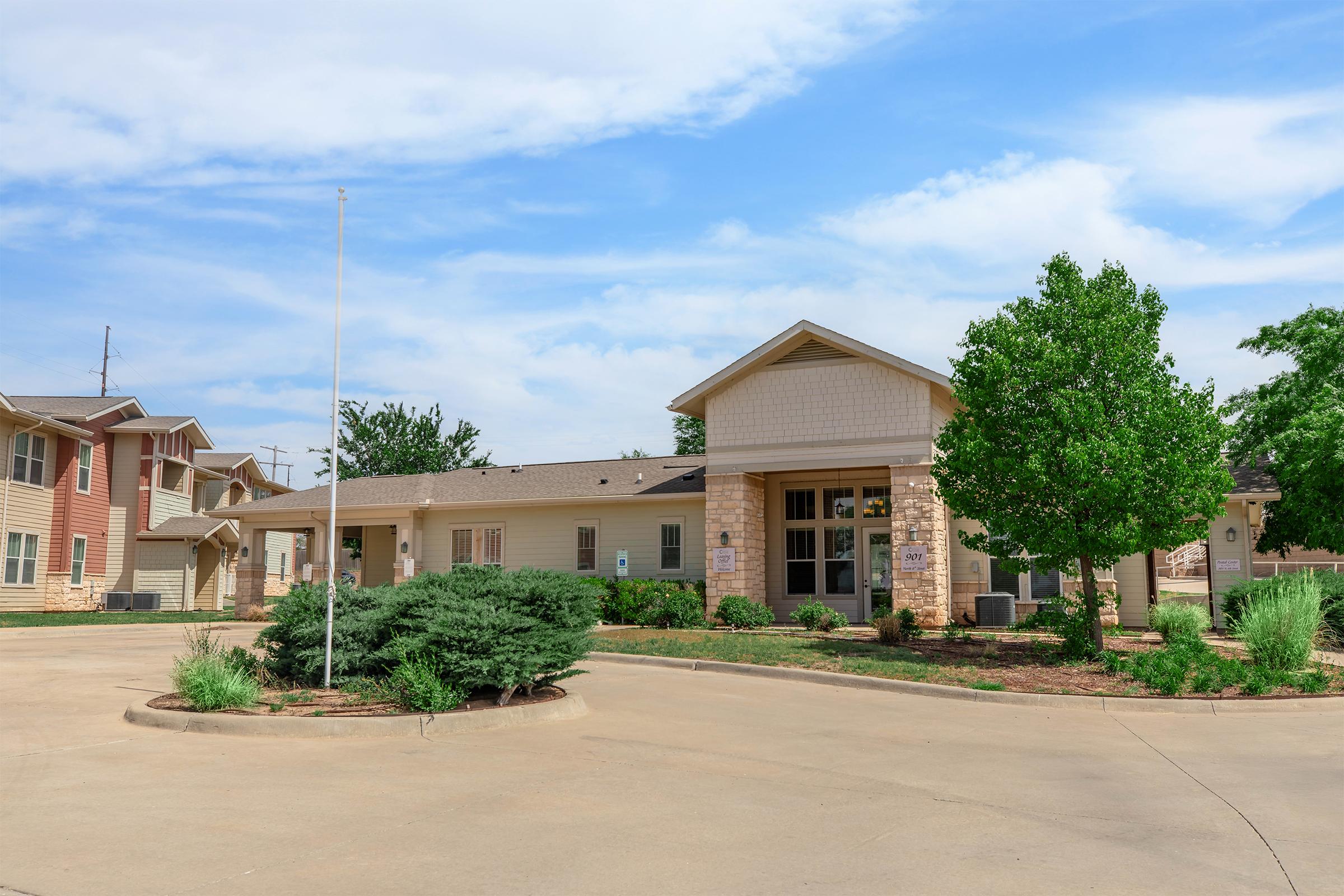 A modern one-story building with a light-colored facade, surrounded by well-maintained landscaping and trees. The entrance features large windows and a covered porch. A circular driveway is visible in front, with a flagpole and greenery enhancing the pleasant appearance of the property under a partly cloudy sky.