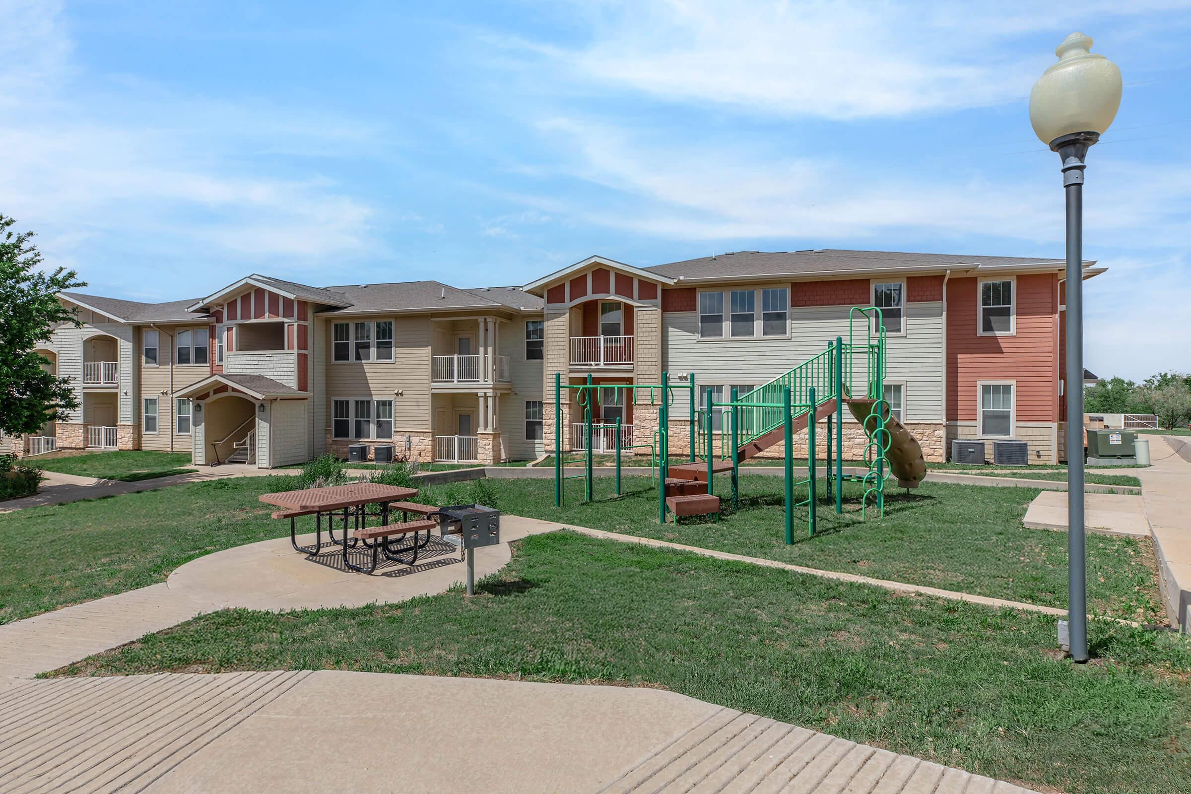 A view of a residential apartment complex featuring two buildings and a playground. The playground includes slides and climbing structures, with picnic tables nearby on the lawn. The area is green and well-maintained, with pathways leading through the property.