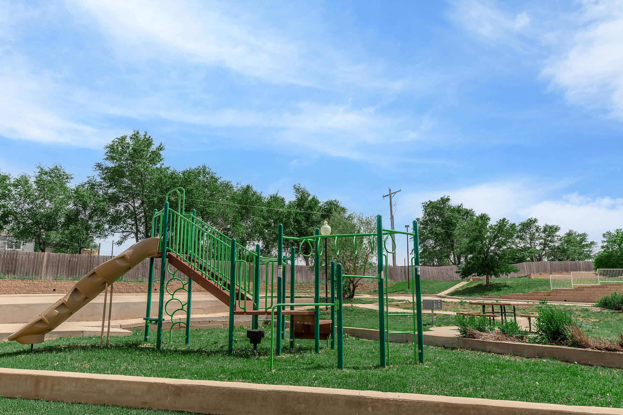A playground featuring a green slide and climbing structure, set in a grassy area. Surrounding the playground are trees and a gently sloping landscape, with picnic tables visible in the background under a clear blue sky.