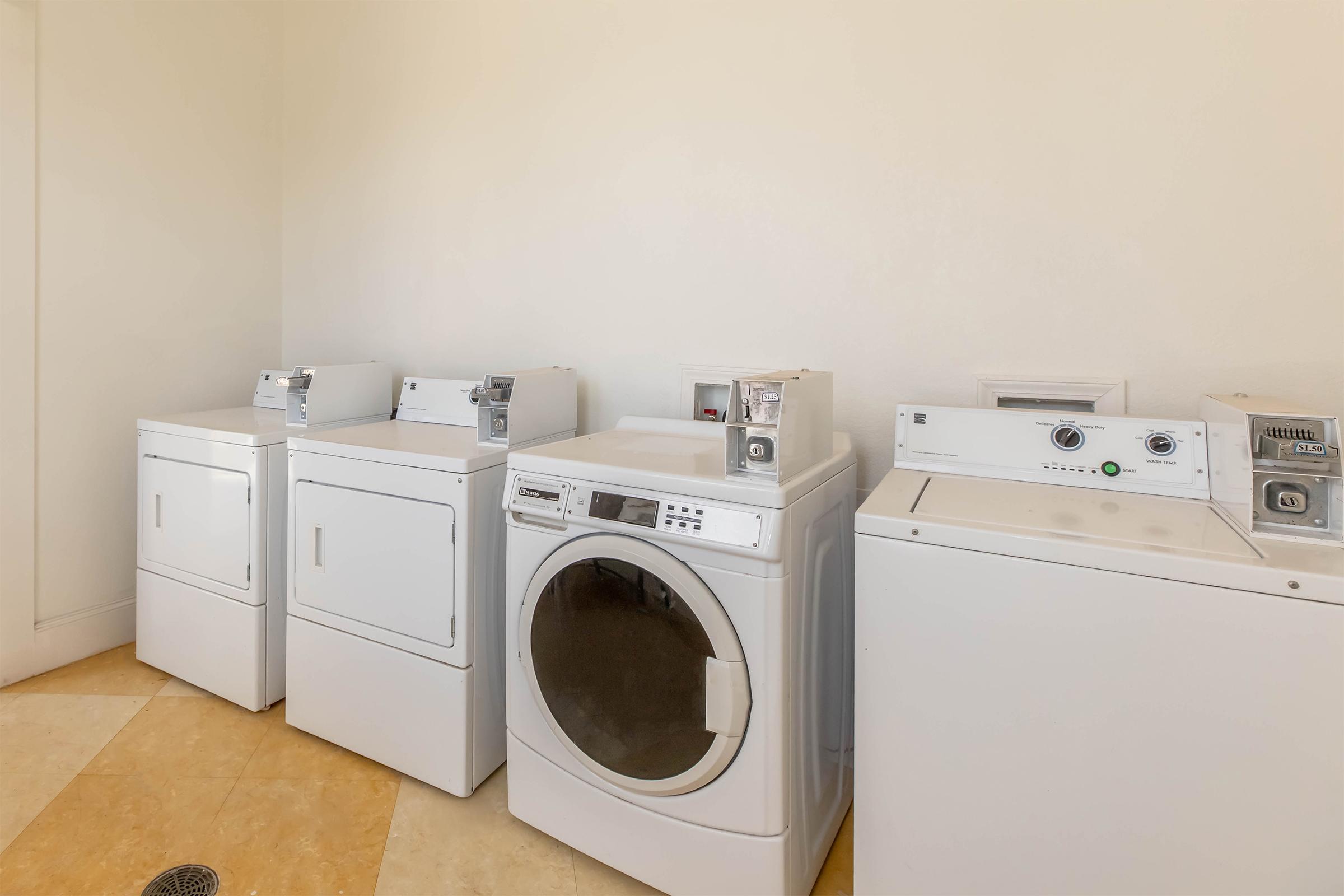 A laundry room featuring four white appliances: two stacked washing machines, a round-front dryer, and a top-loading washing machine. The walls are painted a light color, and the floor is tiled with cream-colored tiles, creating a clean and modern laundry space.