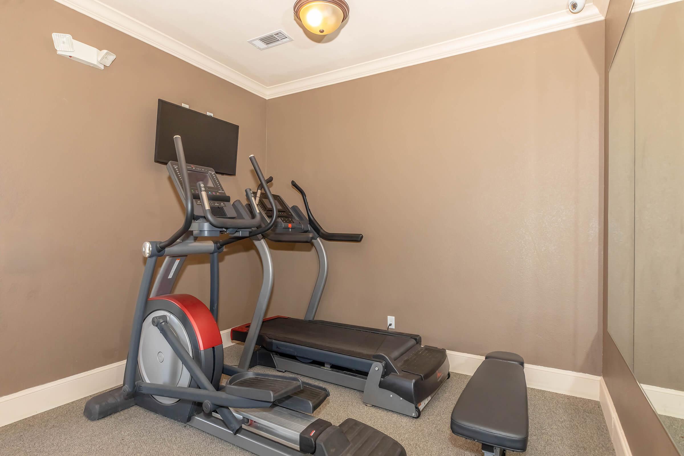 A small home gym featuring a treadmill and an elliptical machine, with a bench for weights. There's a wall-mounted flat-screen TV and a large mirror, all set against a neutral brown wall. The floor is simple, and the ceiling has a light fixture for illumination.
