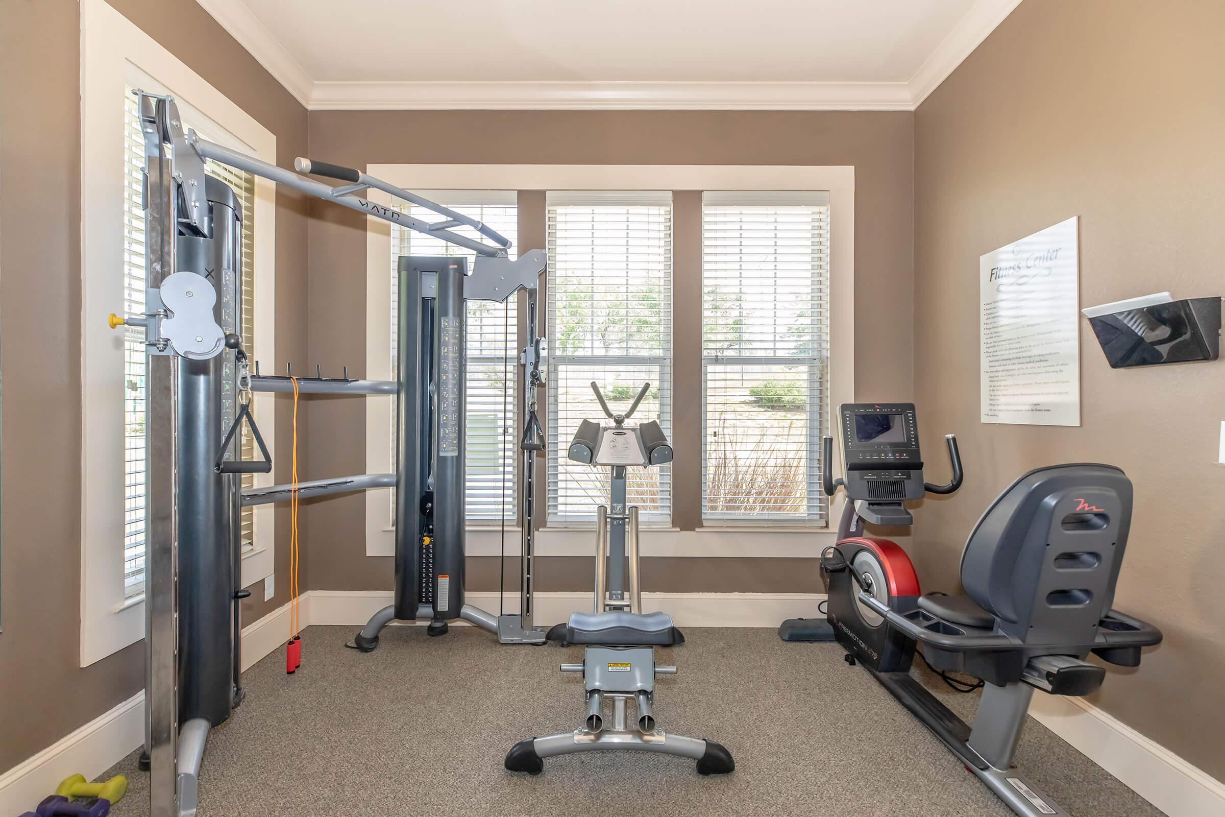 A home gym featuring a multi-function weight machine, a stationary bike, and a cross trainer, positioned in a well-lit room with large windows and neutral-colored walls. The floor is carpeted, and there's a fitness information board on the wall.