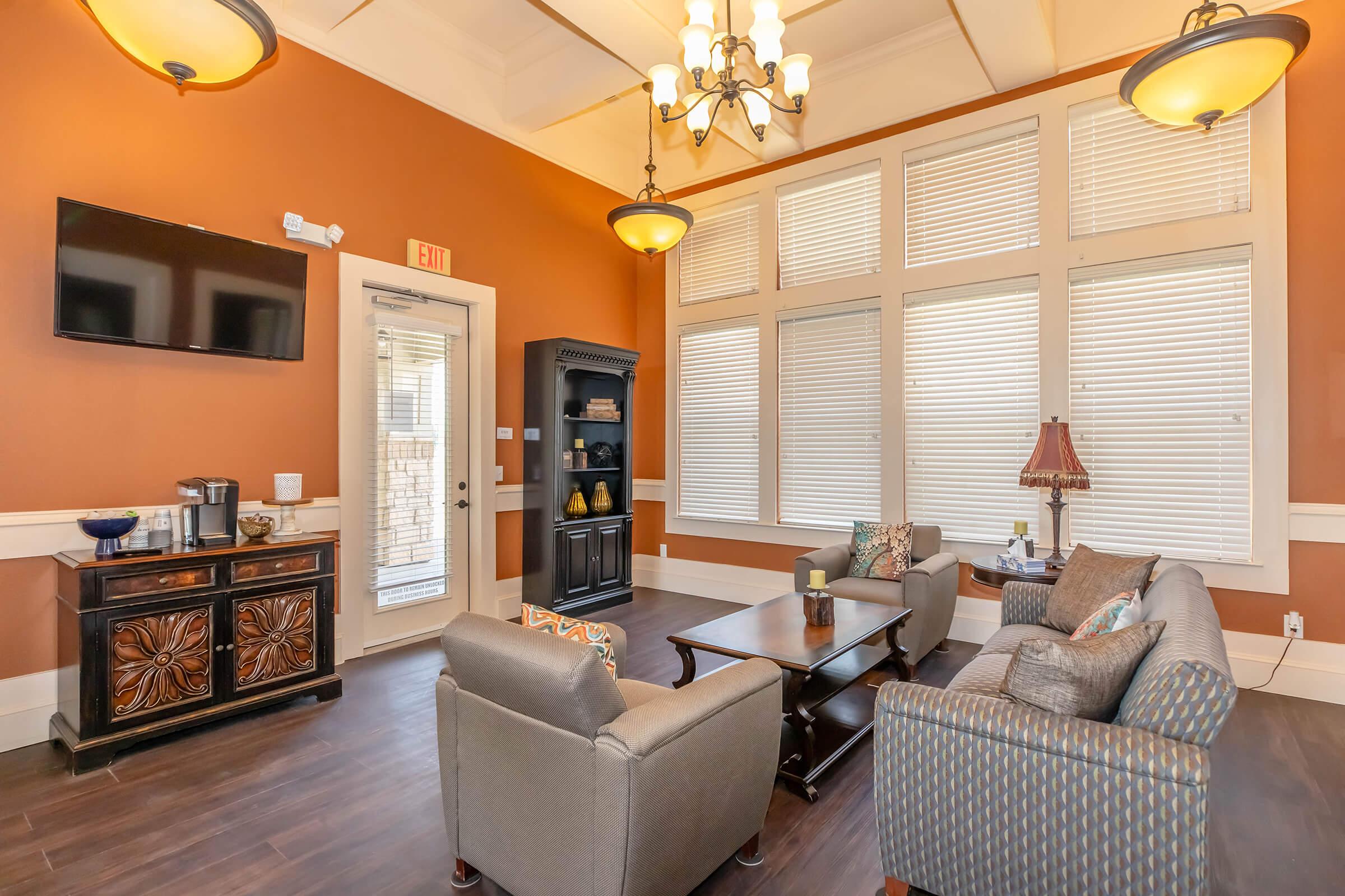 Cozy lounge area featuring a warm orange wall, large windows with blinds, a dark wooden coffee table, two comfortable sofas, an ornate cabinet, and a flat-screen TV. A decorative lamp and coffee station add a welcoming touch, making it a perfect spot for relaxation or socializing.