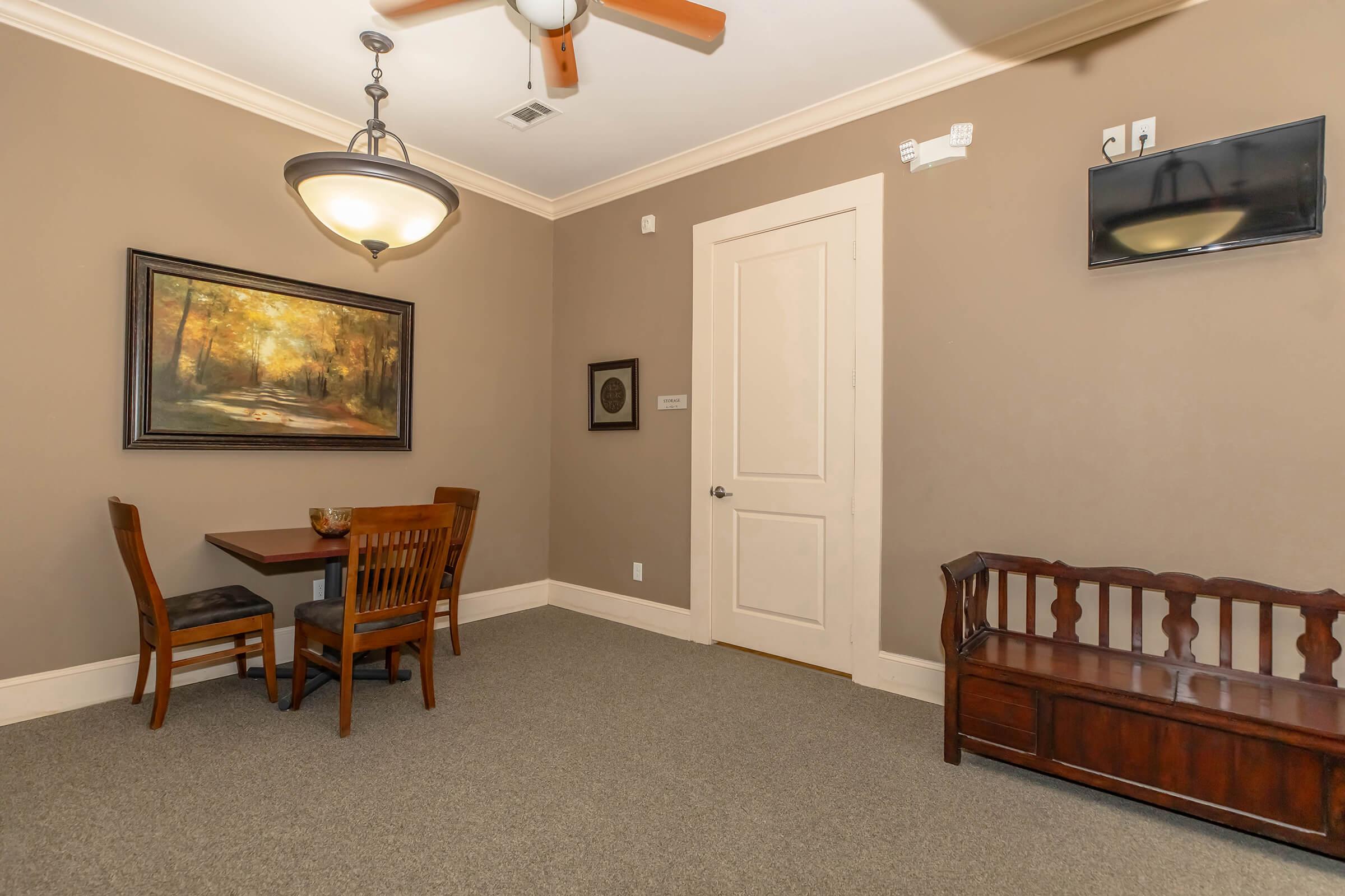 A cozy room featuring a small wooden dining table with two chairs, a decorative painting of a forest scene on the wall, a light fixture, and a wooden bench. A TV is mounted on the wall, and the floor is carpeted. The walls are painted in a warm, neutral tone.