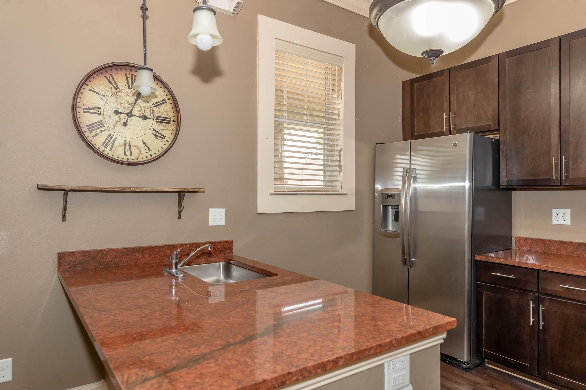 A modern kitchen featuring dark wooden cabinets, a stainless steel refrigerator, a sink with a granite countertop, and a large wall clock. Natural light enters through a window, illuminating the space with warm tones of brown and red. Decorative shelf is mounted on the wall.