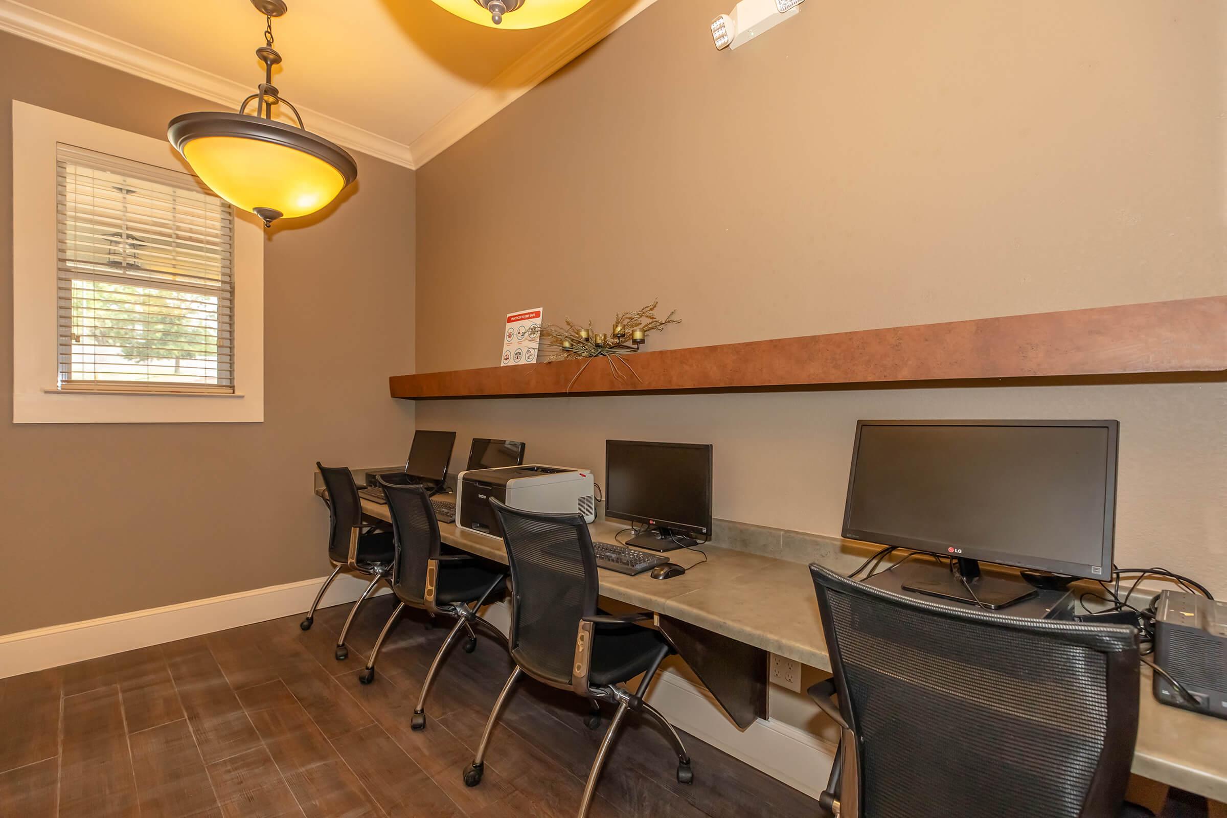 A small office space featuring four black chairs positioned at a long gray desk with computer monitors. The walls are painted a warm brown, and overhead, there are pendant lights. A decorative shelf with minimal decor is mounted above the desk, and a window allows natural light to enter the room.