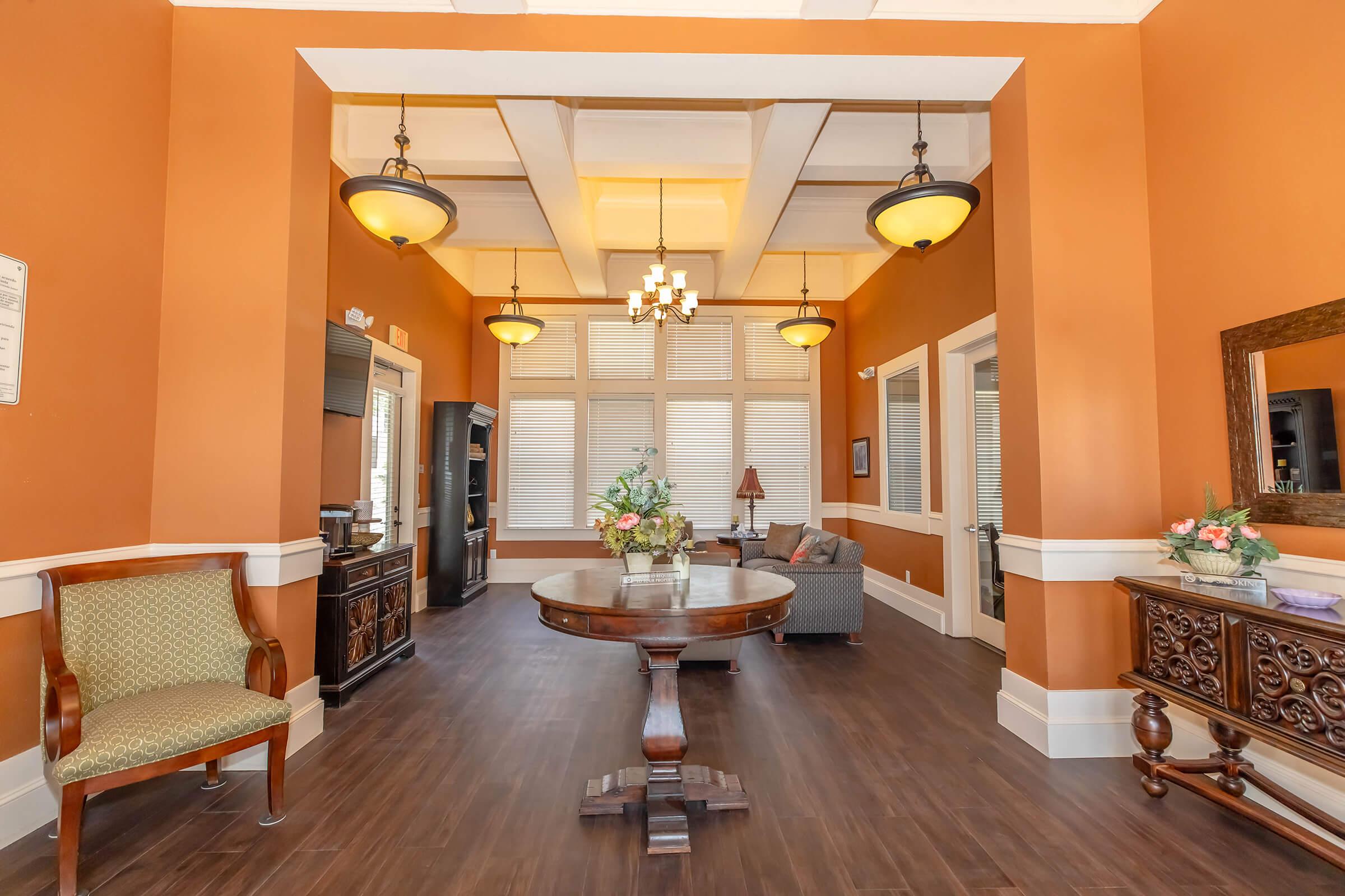 A cozy interior room featuring an inviting seating area with a grey sofa, a wooden table at the center, and elegant lighting fixtures. The walls are painted in warm orange tones, complemented by decorative furniture, including a vintage side table and a chair. Large windows allow natural light to brighten the space.