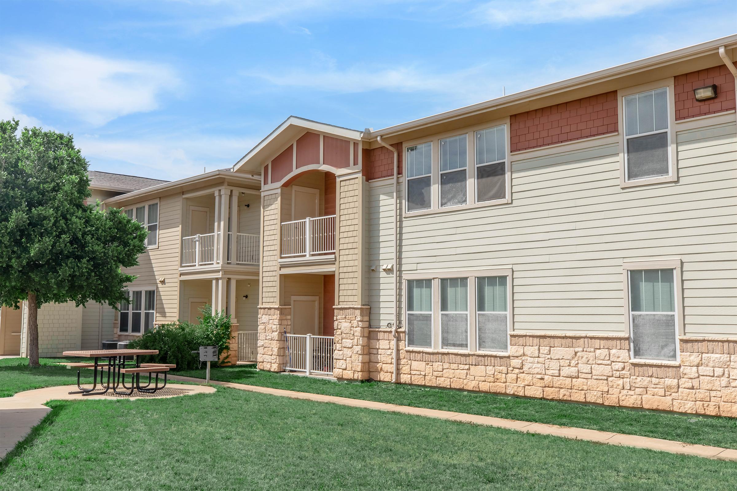 A two-story residential building with beige siding and stone accents, featuring balconies on the upper level. A small courtyard with a round picnic table is visible on the green lawn in front of the building. The sky is clear with a few clouds, creating a bright and inviting atmosphere.