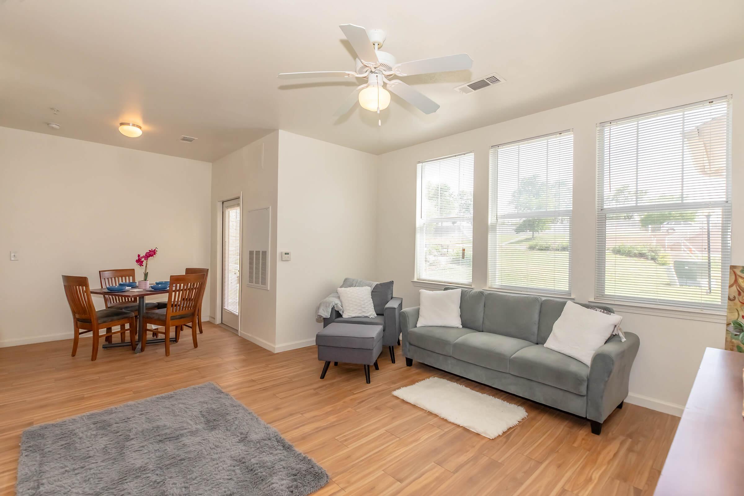 Bright and spacious living room featuring a gray couch with white pillows, a light rug, and a ceiling fan. A dining area with a wooden table and chairs is visible in the background. Large windows allow natural light to fill the space, creating a warm and inviting atmosphere.