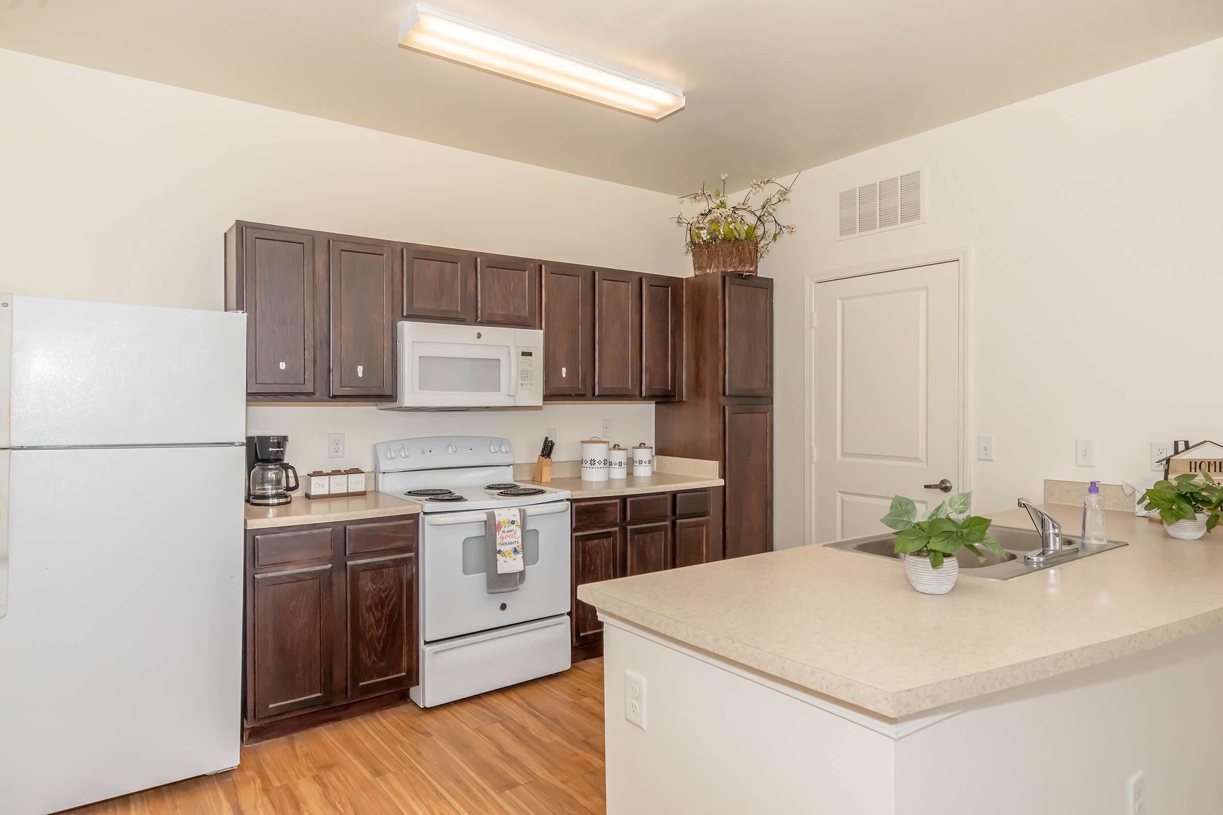 Modern kitchen featuring dark wood cabinets, a white refrigerator, and a white stove with a microwave above. A countertop with a sink and a small potted plant is visible. The kitchen has warm wooden flooring and light walls, creating a bright and inviting atmosphere.