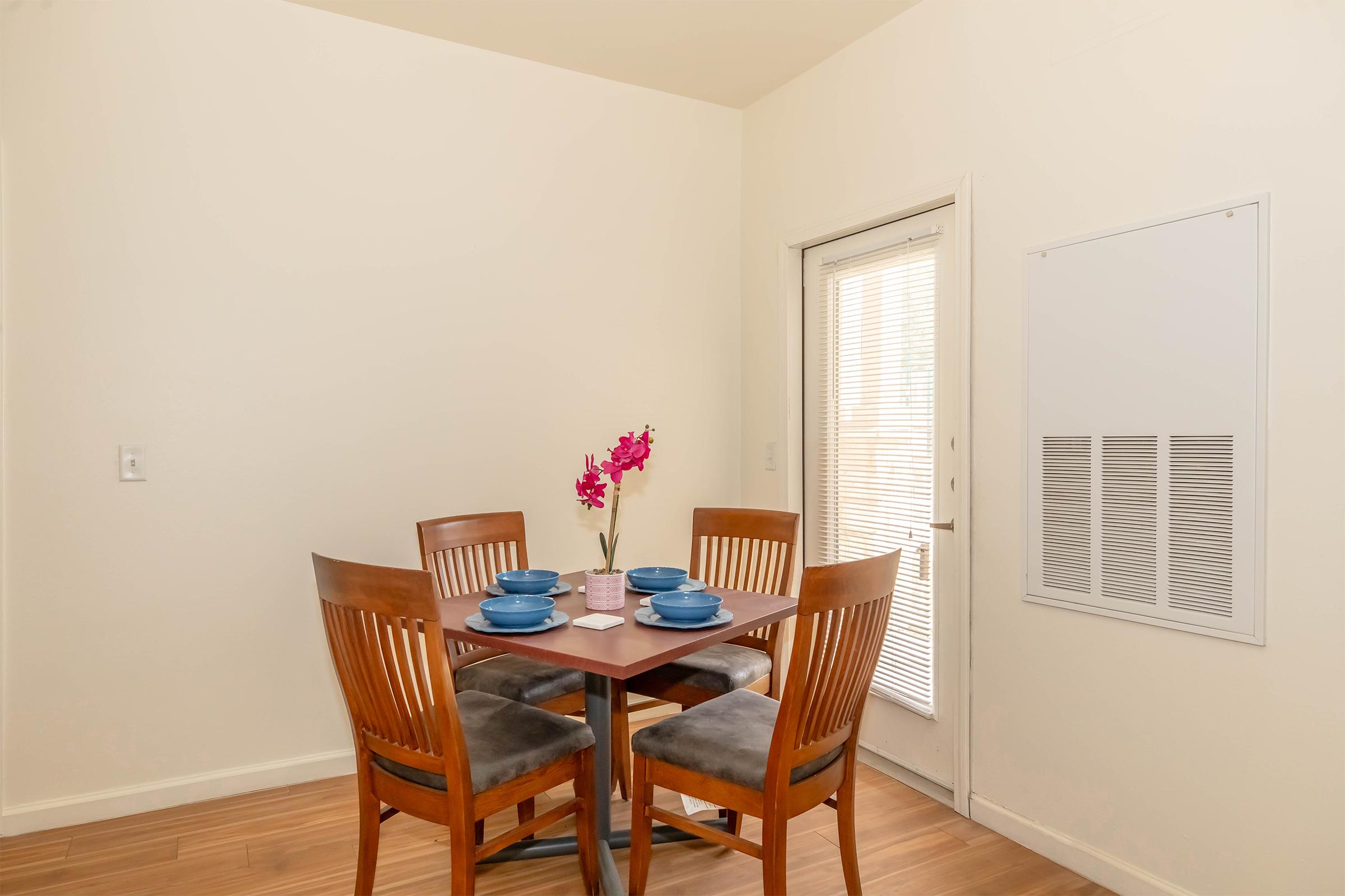 A small dining area featuring a square table set with blue plates and a pink flower arrangement. Four wooden chairs surround the table. A sliding glass door leads to the outside, and a wall vent is visible nearby. The walls are painted a light color, creating a bright, inviting atmosphere.