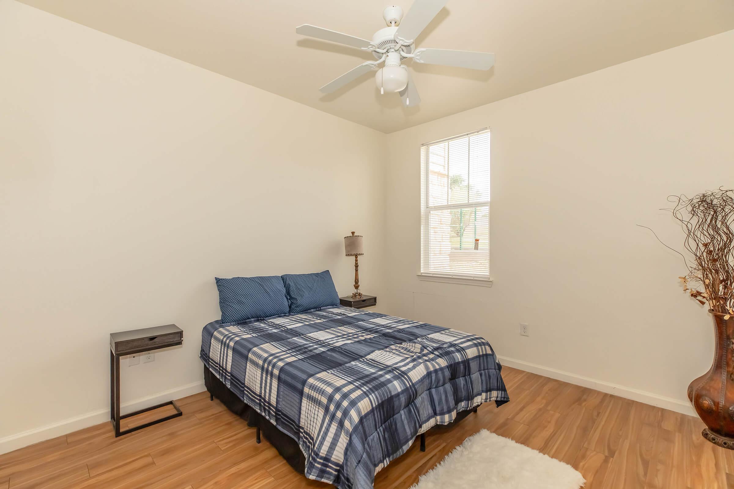 A simple bedroom featuring a bed with a blue plaid comforter, a nightstand with a lamp, a rug on the wooden floor, and a window allowing natural light. The walls are painted white, and a ceiling fan is installed.