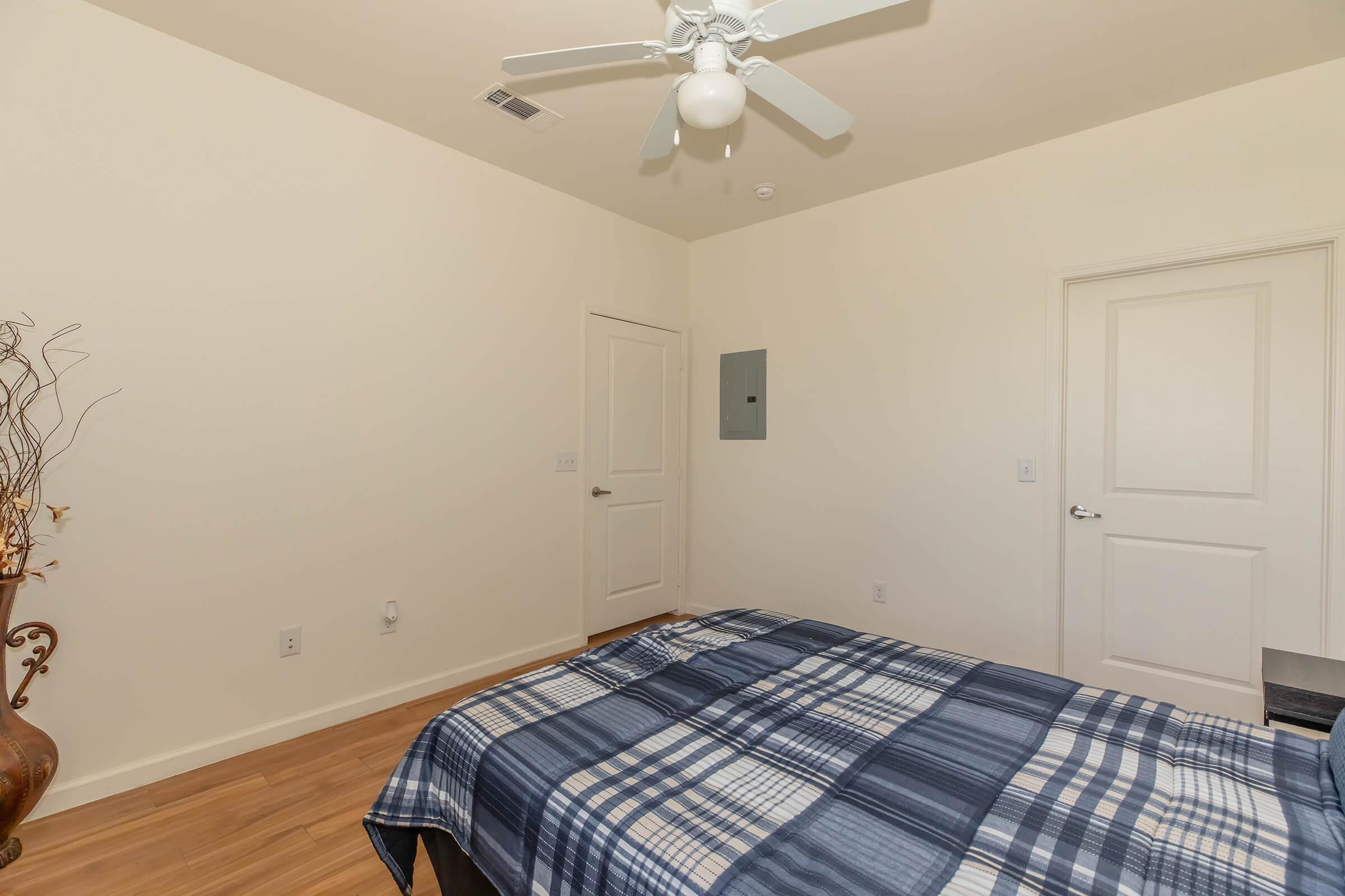 A simple bedroom featuring a neatly made bed with a blue and white plaid blanket. There is a light-colored wall, a ceiling fan, and a closed door. A decorative plant is placed in the corner, and an electrical panel is mounted on the wall. The flooring is wooden.