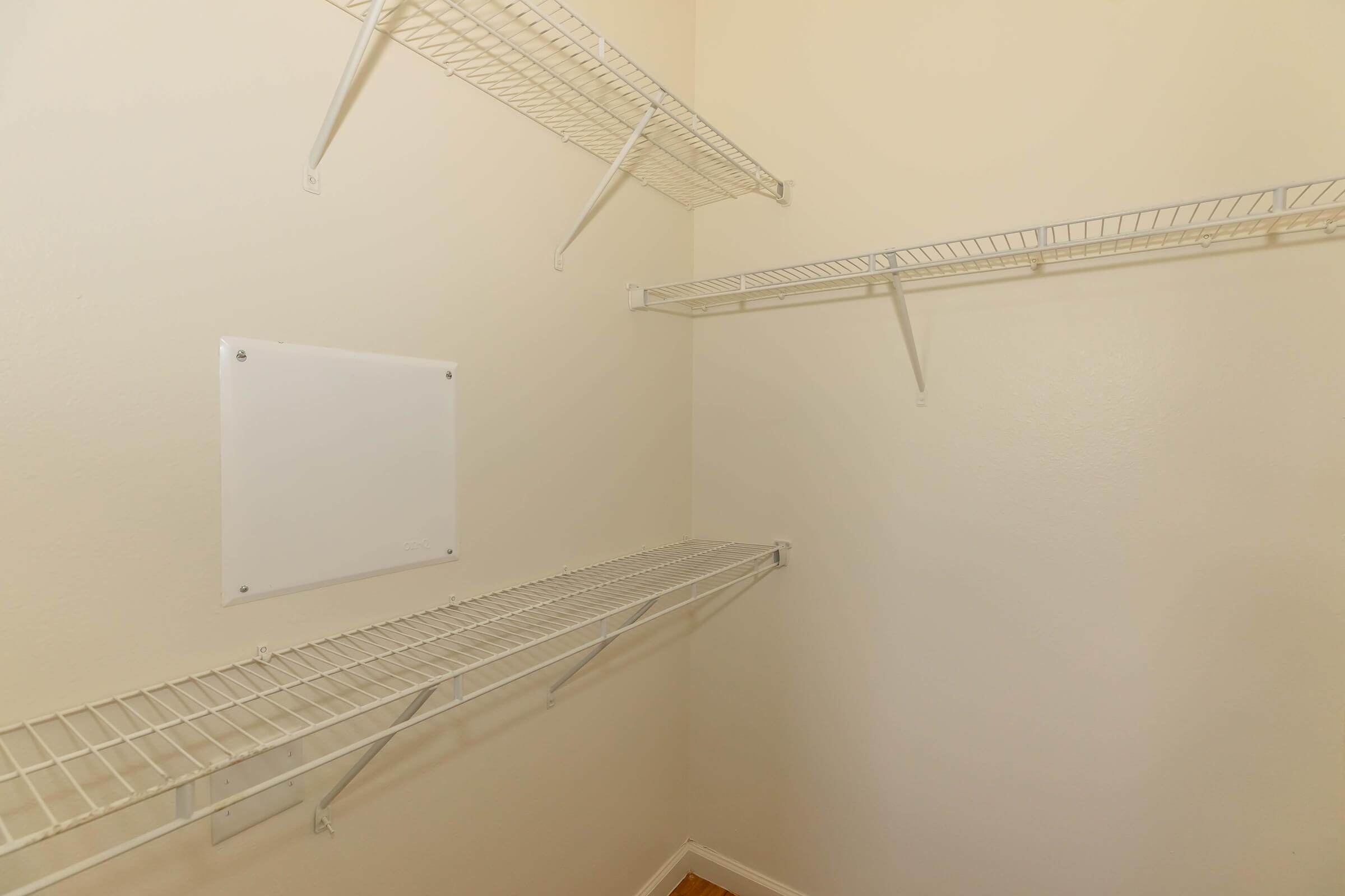 A clean, empty closet with light-colored walls and white wire shelving. The closet features two horizontal shelves and one vertical panel on the wall, providing storage space. The floor is hardwood, adding warmth to the otherwise minimalist design.