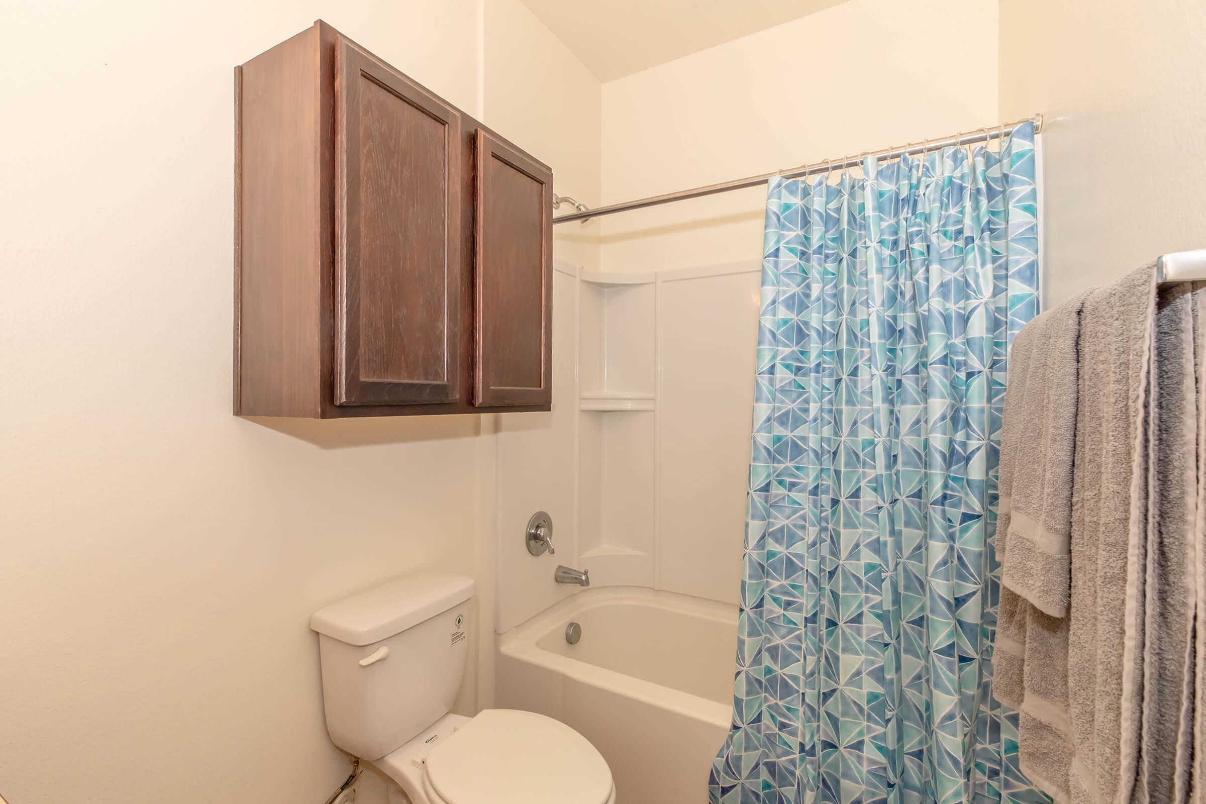 A small bathroom featuring a bathtub with a blue geometric shower curtain, a white toilet, and a dark wood cabinet above. Towels hang on a rack, and the walls are painted in a light color, creating a bright and clean appearance.