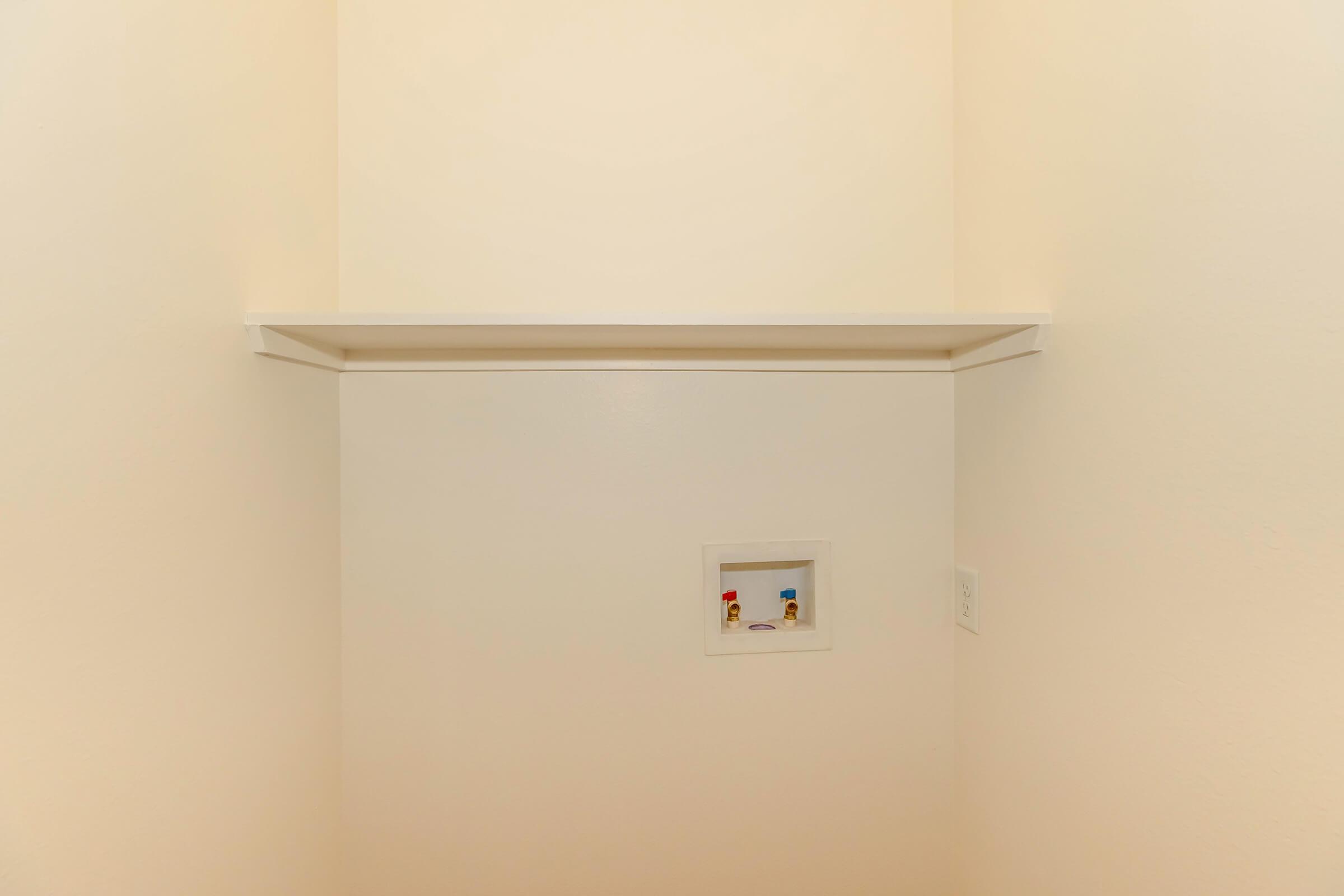 A clean corner of a room featuring a white shelf mounted on the wall. Below the shelf, there is a small recessed area with two colorful figurines displayed inside. The walls are painted a light color, creating a bright and minimalistic aesthetic.