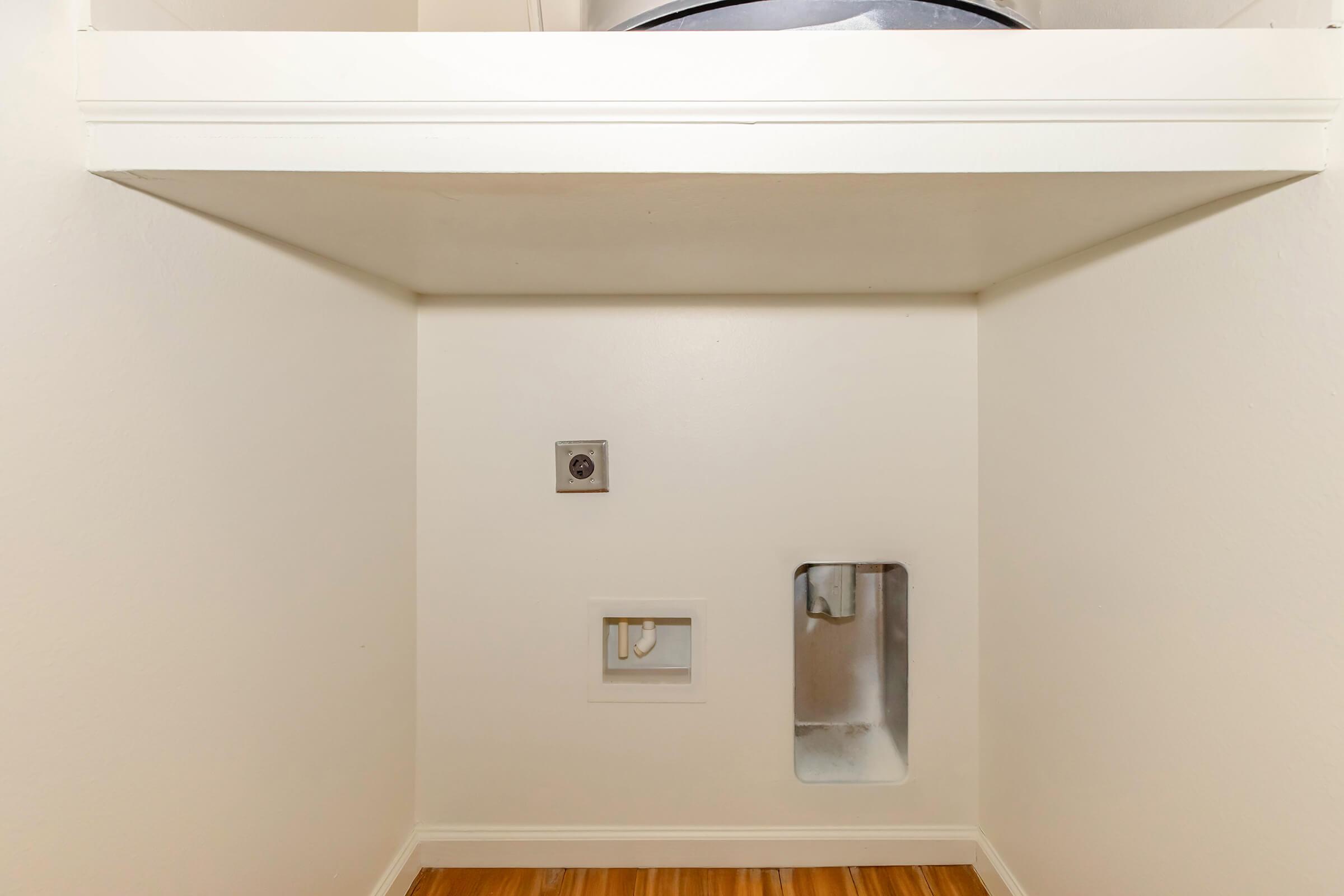 Empty laundry room space featuring a white wall with a shelf above. There are two utility openings on the wall: one for a dryer vent and another for plumbing connections. The floor has wood-like laminate, and the overall area is clean and uncluttered, showcasing a simple, functional design.
