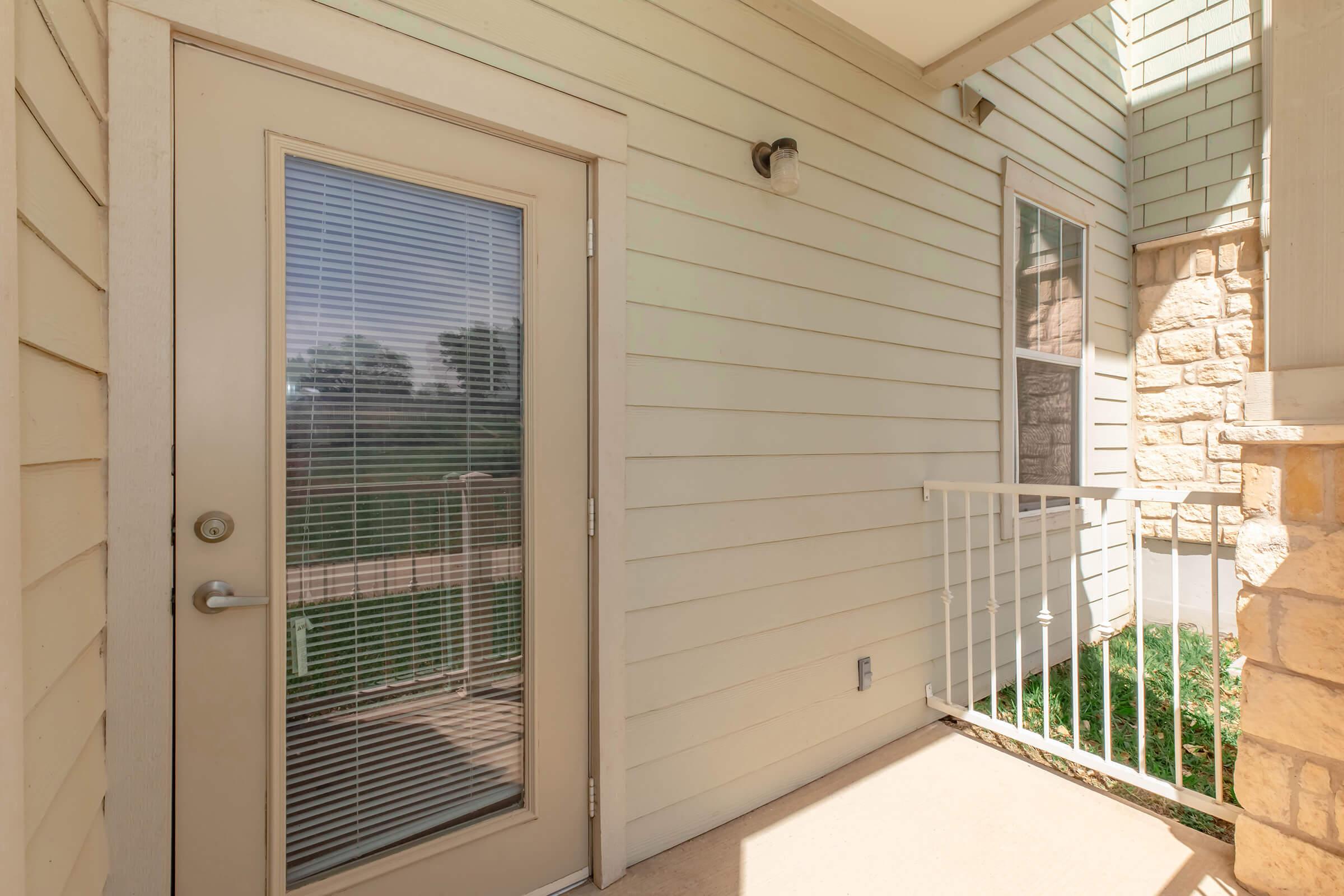 A close-up view of a light-colored door with a vertical window, located on a beige exterior wall of a building. The surrounding area features a small railing and well-maintained grass, indicating an outdoor entrance to a residential space. Natural light casts shadows on the surface.