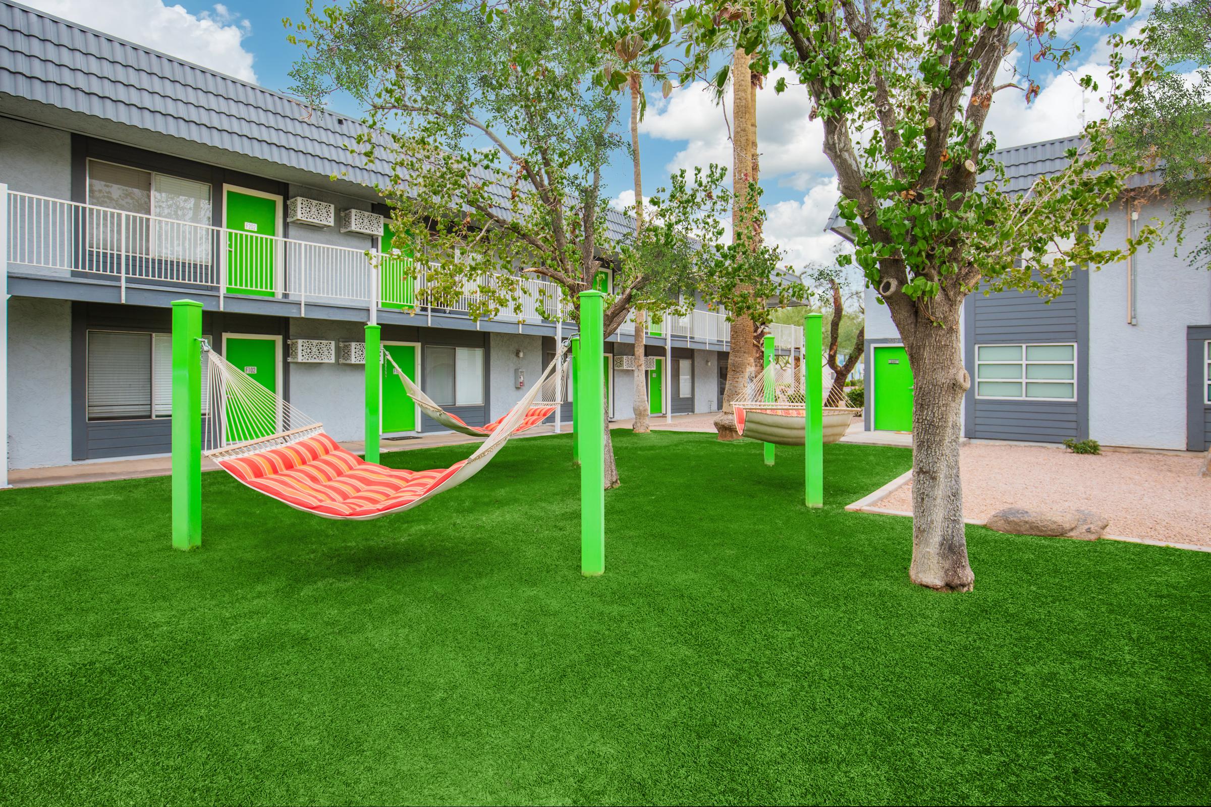 A vibrant courtyard featuring two colorful hammocks suspended between green poles on a lush, artificial lawn. Surrounding the area are modern buildings with gray exteriors and bright green doors. Trees provide some shade under a partly cloudy sky.