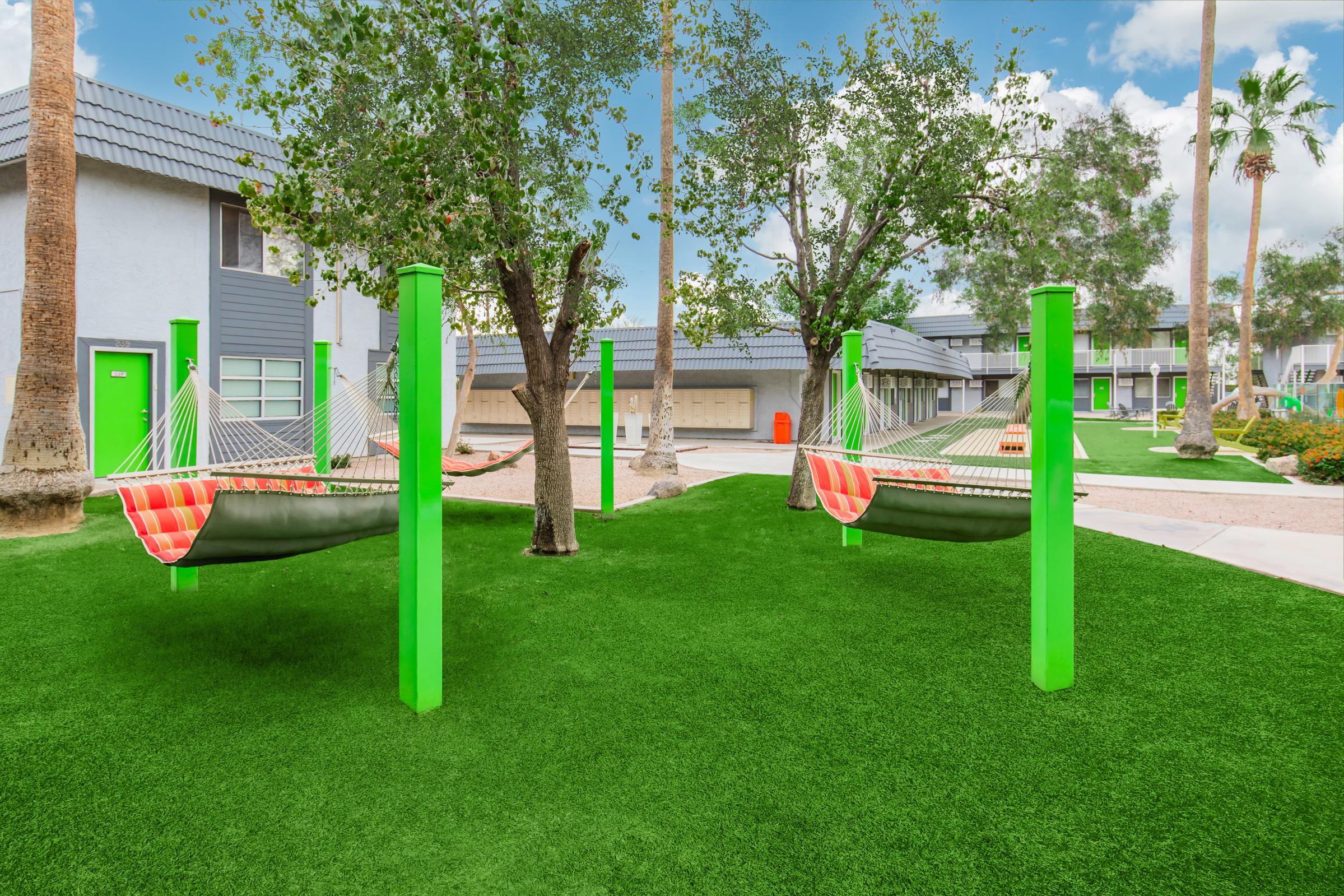 Two colorful hammocks are suspended between bright green posts in a well-maintained outdoor area. Lush artificial grass covers the ground, surrounded by trees and modern buildings. The scene exudes a relaxing atmosphere, perfect for lounging on a sunny day.