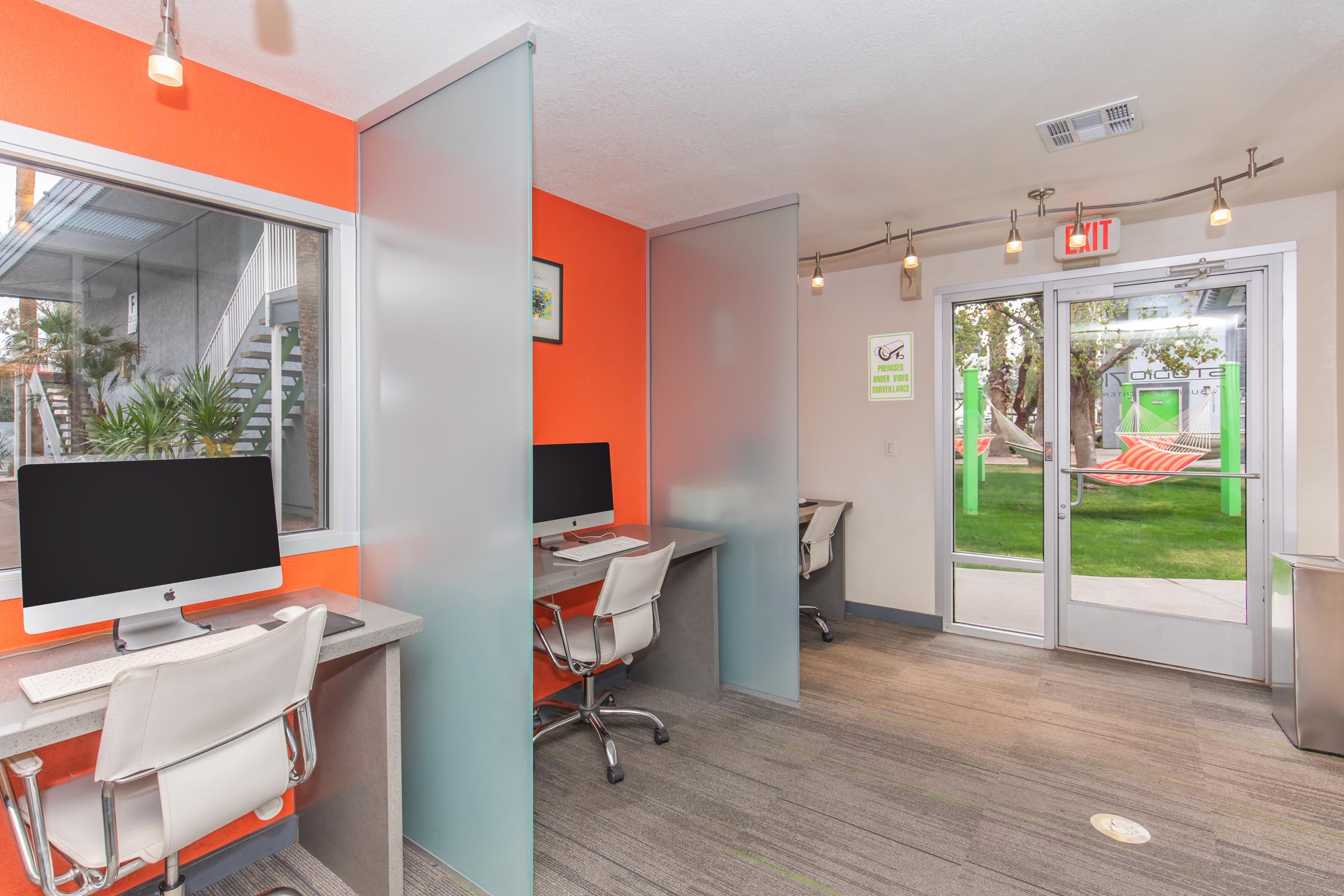 Modern office space featuring three workstations with desktop computers against a bright orange wall. Frosted glass partitions separate the areas. A door leads outside, revealing a green hammock and patio area. The workspace is well-lit with overhead pendant lights and has a stylish, professional look.