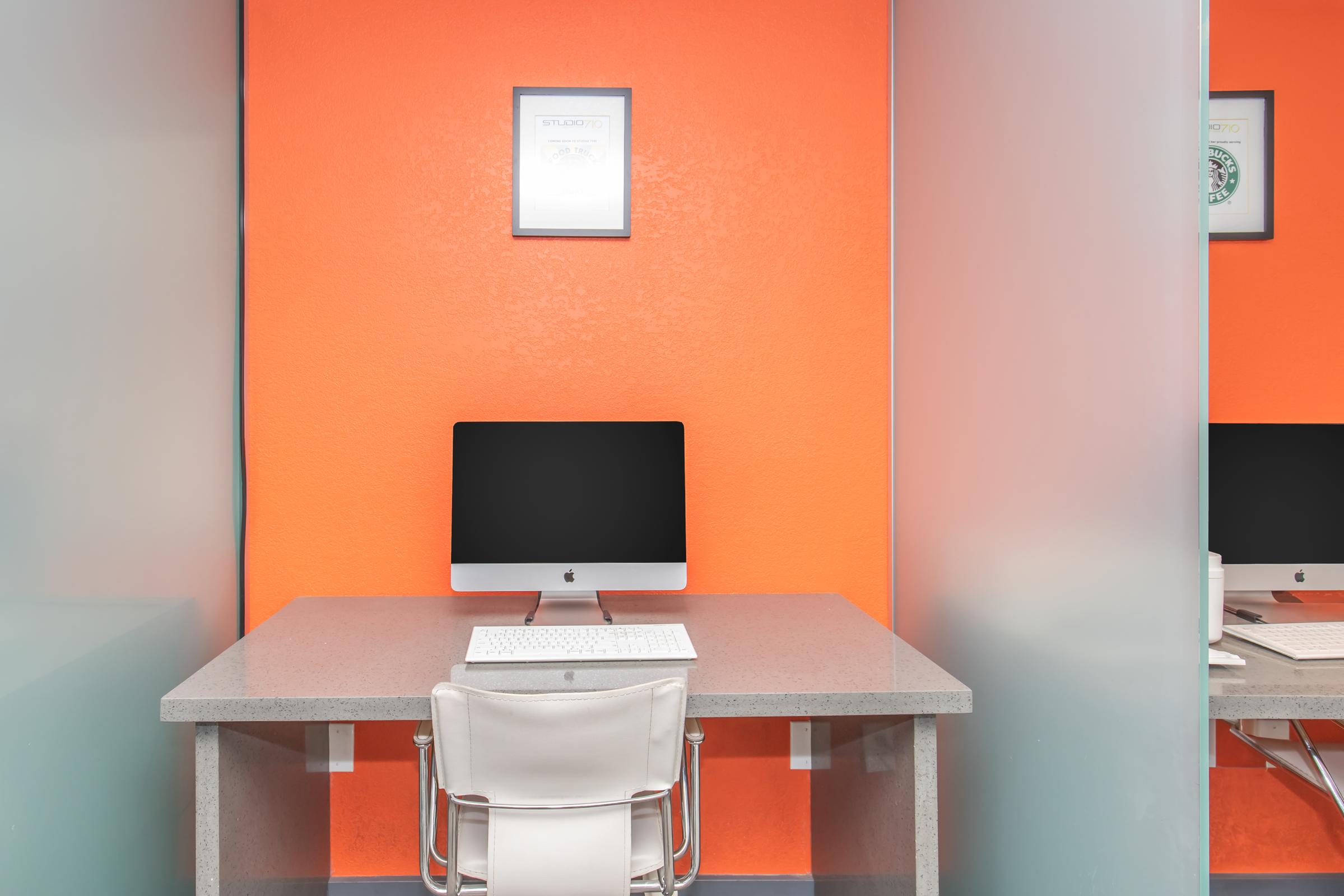 A modern workspace featuring a sleek desk with an iMac computer and a keyboard. The backdrop is a vibrant orange wall, and there's a framed picture above the computer. A clear partition on the side provides a sense of privacy, while a white chair completes the minimalist design.