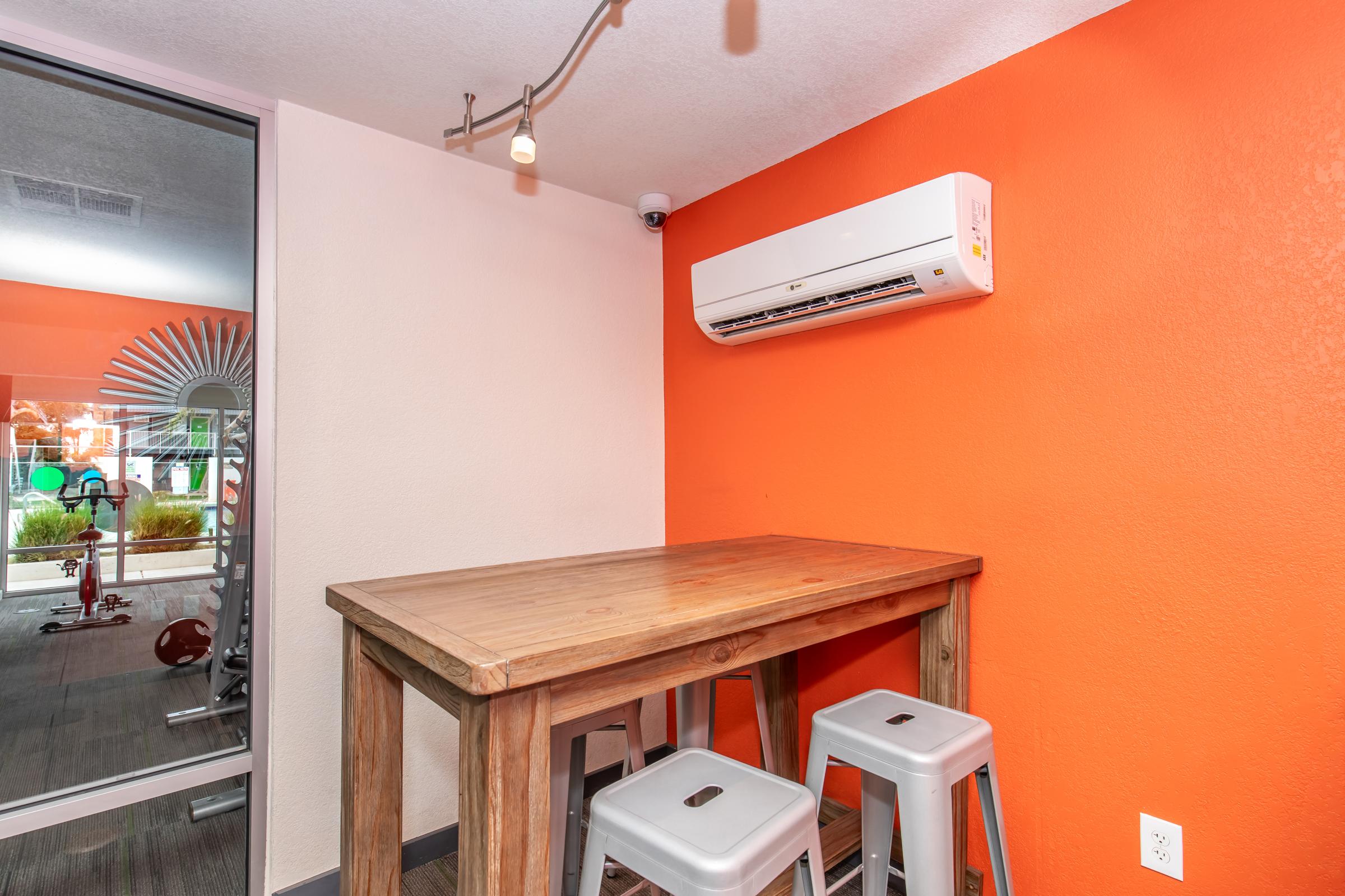 A corner of a modern gym or lounge featuring a wooden table with three metal stools, an air conditioning unit on the wall, and an orange accent wall. Behind, a glimpse of workout equipment and greenery is visible, suggesting a fitness-focused environment.