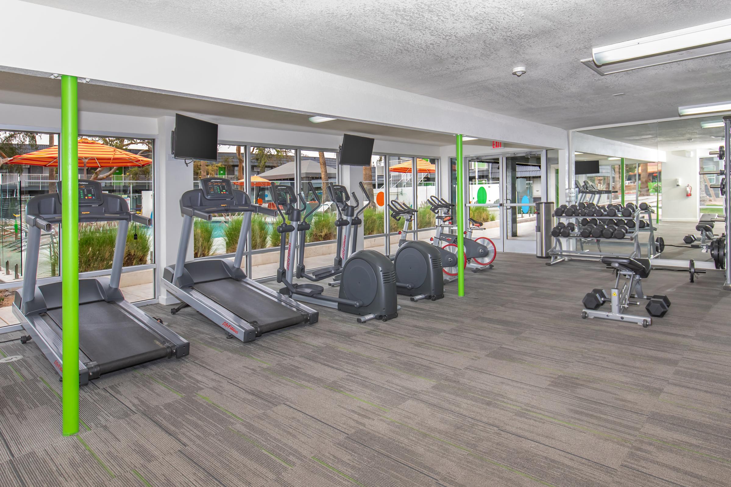 A modern gym featuring multiple treadmills, ellipticals, and a set of free weights. Large windows provide natural light and overlook a pool area with orange umbrellas. The flooring is carpeted in neutral tones, and there are two flat-screen TVs mounted on the walls.