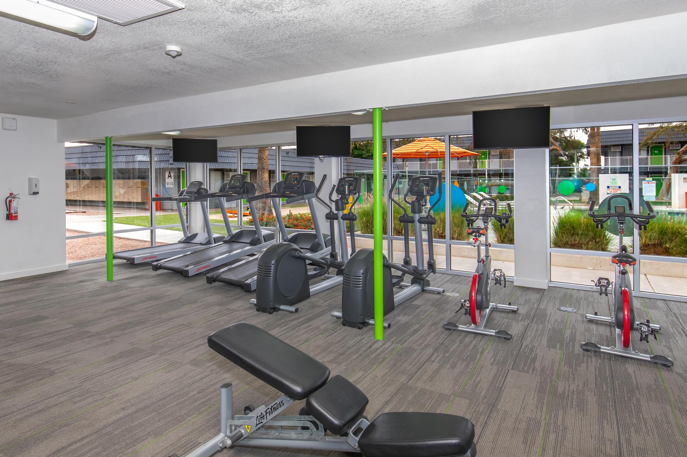 A modern gym with cardio machines such as treadmills and stationary bikes. Large windows provide natural light and a view of an outdoor area with a pool and lounge chairs. The room features green accents and multiple flat-screen TVs mounted on the walls. Fitness equipment is arranged neatly on a carpeted floor.