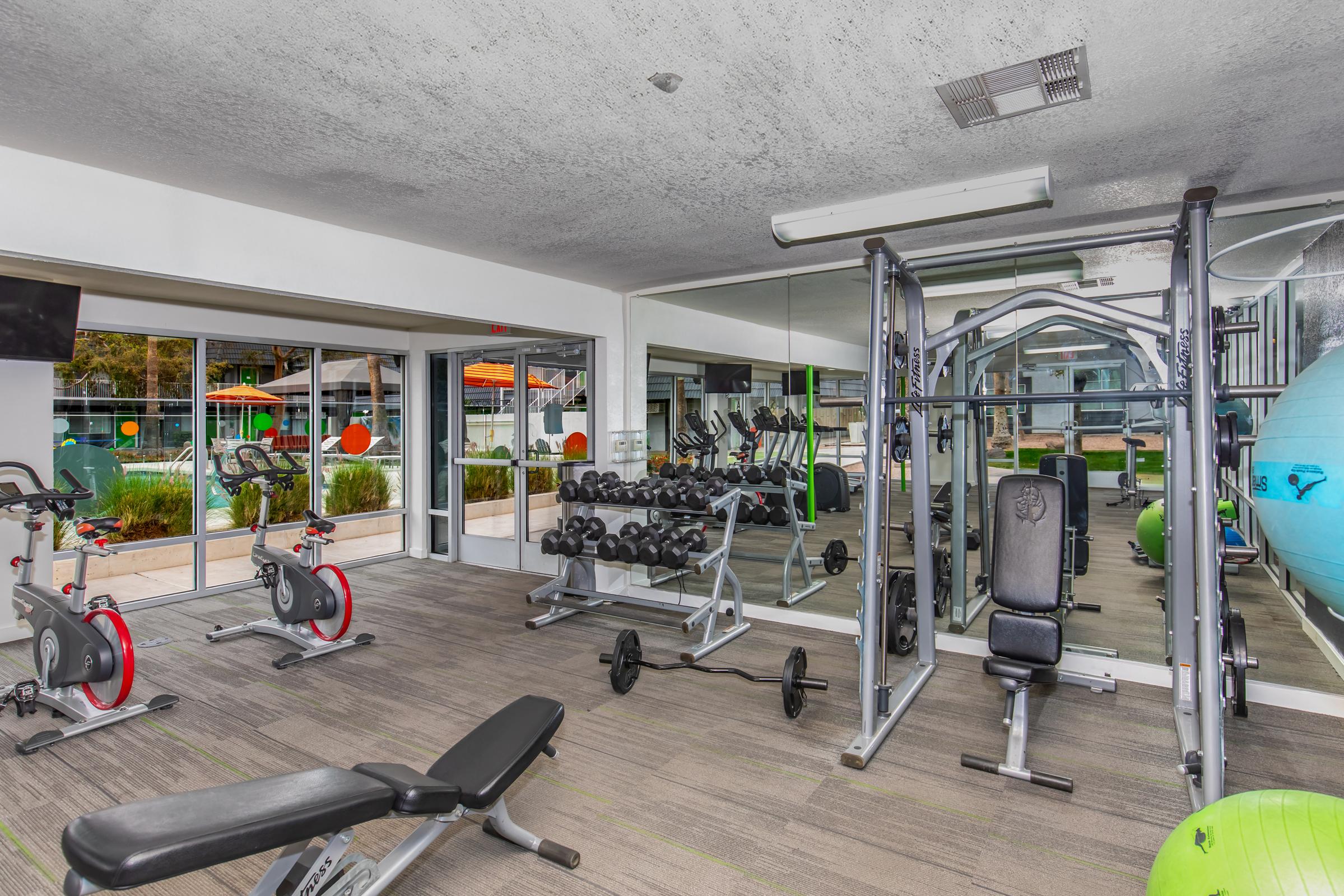 A modern gym interior featuring various workout equipment, including weight benches, free weights, stationary bikes, and a cable machine. Large windows provide natural light and a view of an outdoor pool area with lounge chairs and vibrant green accents. Mirrors line one wall, enhancing the feeling of space.