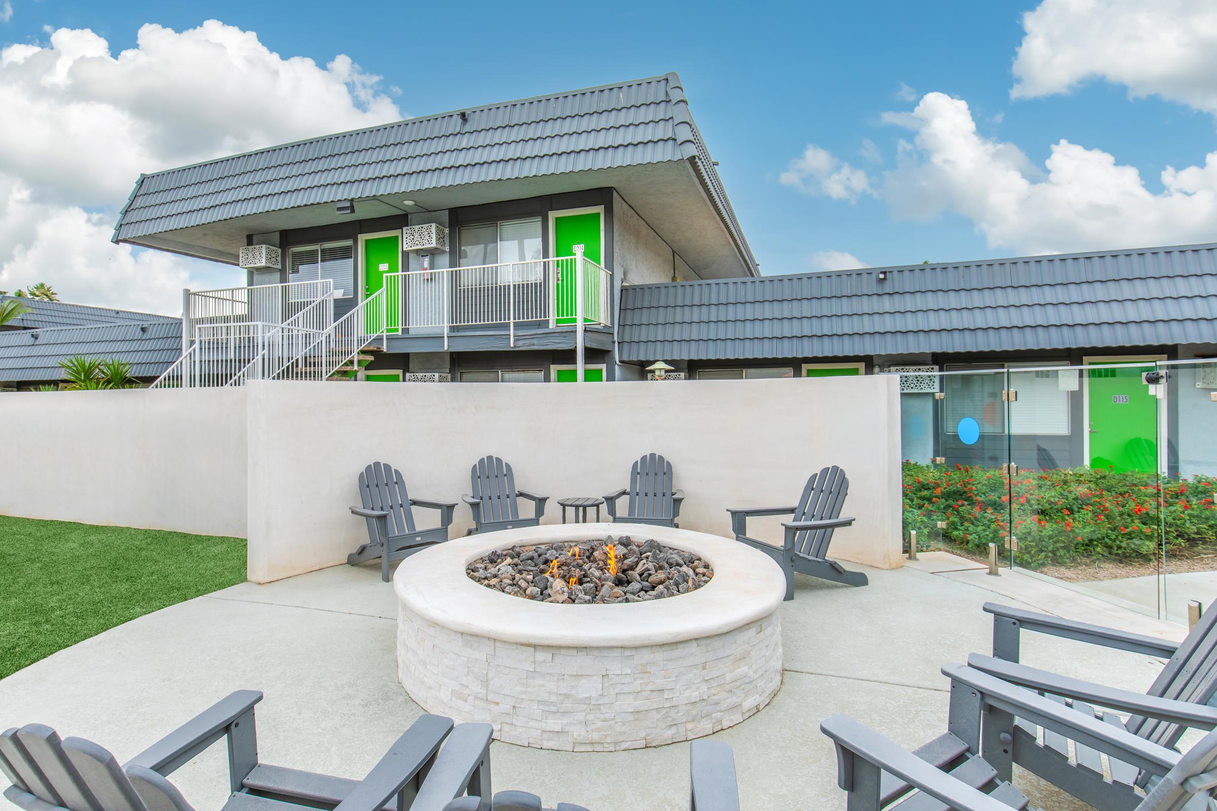 A modern outdoor space featuring a circular fire pit surrounded by gray adirondack chairs. In the background, a two-story building with a gray roof and green doors. Lush landscaping and blue skies complete the inviting atmosphere.