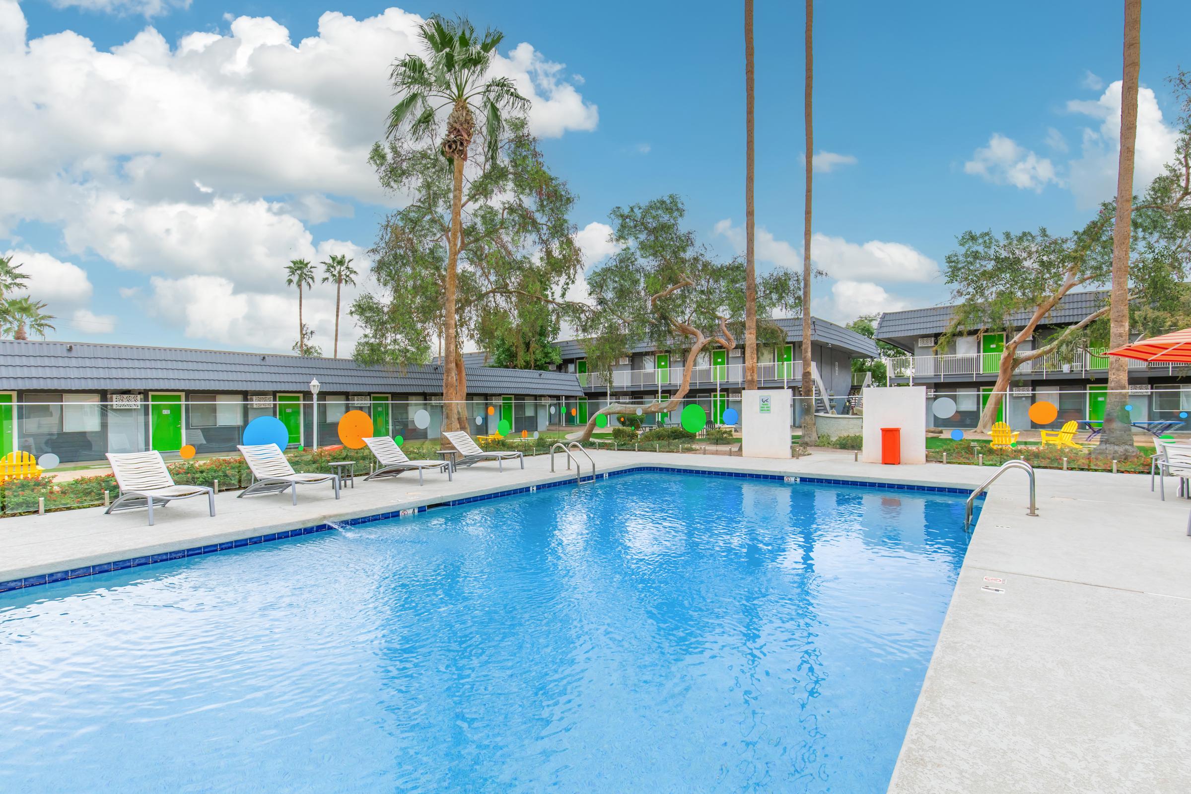 A bright, inviting outdoor pool area surrounded by palm trees, with lounge chairs arranged along the water's edge. Colorful circular decorations are seen on the walls of nearby buildings, and the sky is clear with some clouds, creating a vibrant, relaxing atmosphere.