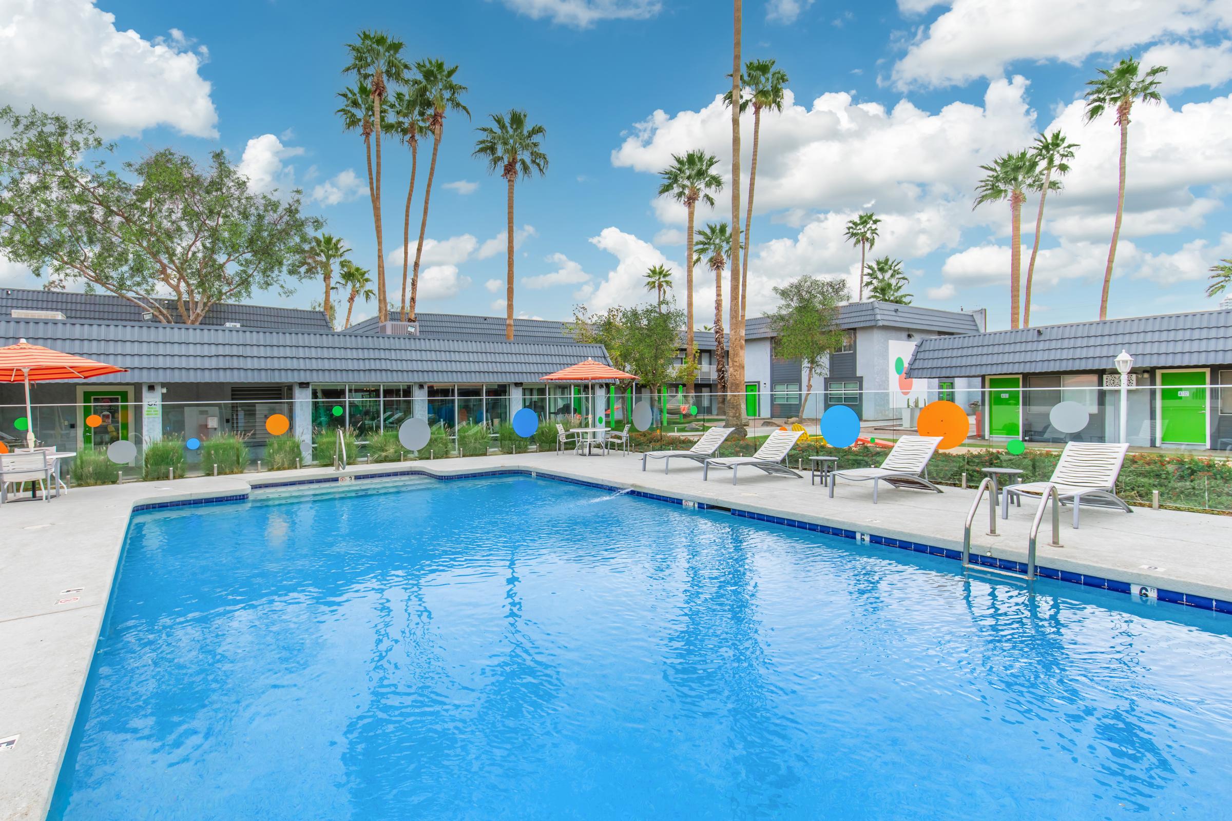 Swimming pool with clear blue water surrounded by lounge chairs and colorful umbrellas. Palm trees in the background complement the sunny, blue sky with fluffy white clouds. Modern buildings with bright accents are adjacent to the pool area, creating a vibrant and inviting atmosphere.