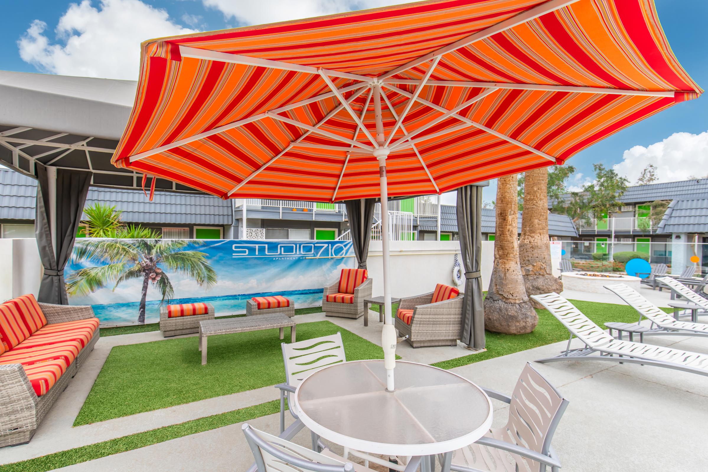 A bright outdoor lounge area featuring a striped orange and white umbrella over a round table, surrounded by wicker chairs and loungers. In the background, a vibrant mural of a beach scene can be seen, along with palm trees and a well-maintained grassy area, creating a relaxing atmosphere.