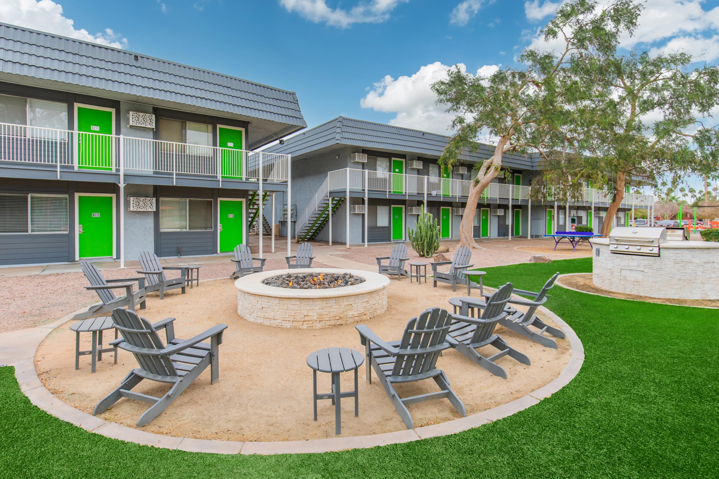 A modern outdoor space with a circular fire pit surrounded by gray Adirondack chairs. Nearby, a grassy area and a barbecue station are visible, along with two-story apartment buildings featuring gray exteriors and bright green doors. The sky is partly cloudy, creating a welcoming atmosphere.