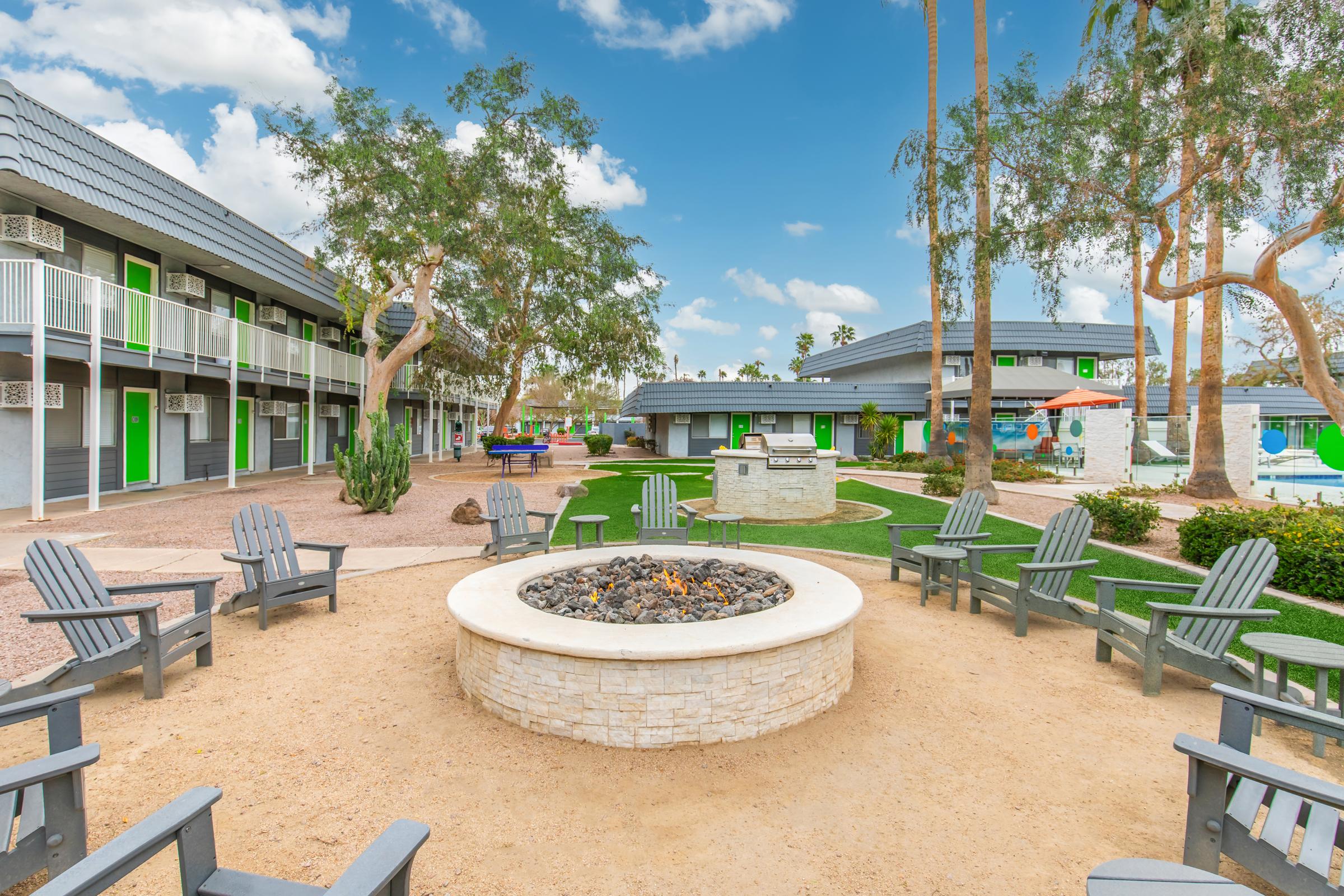 A cozy outdoor area featuring a circular stone fire pit surrounded by wooden seating. Lush green grass, palm trees, and a colorful poolside area are visible in the background, with modern buildings housing guest rooms with green accents under a partly cloudy sky.