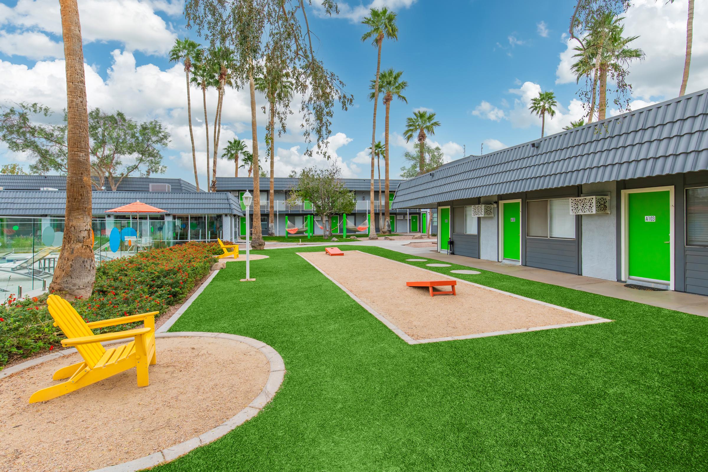 A vibrant courtyard scene featuring artificial grass, lounge chairs, and palm trees. The layout includes a sand court, picnic benches, and colorful green doors on nearby buildings under a bright blue sky with fluffy clouds.