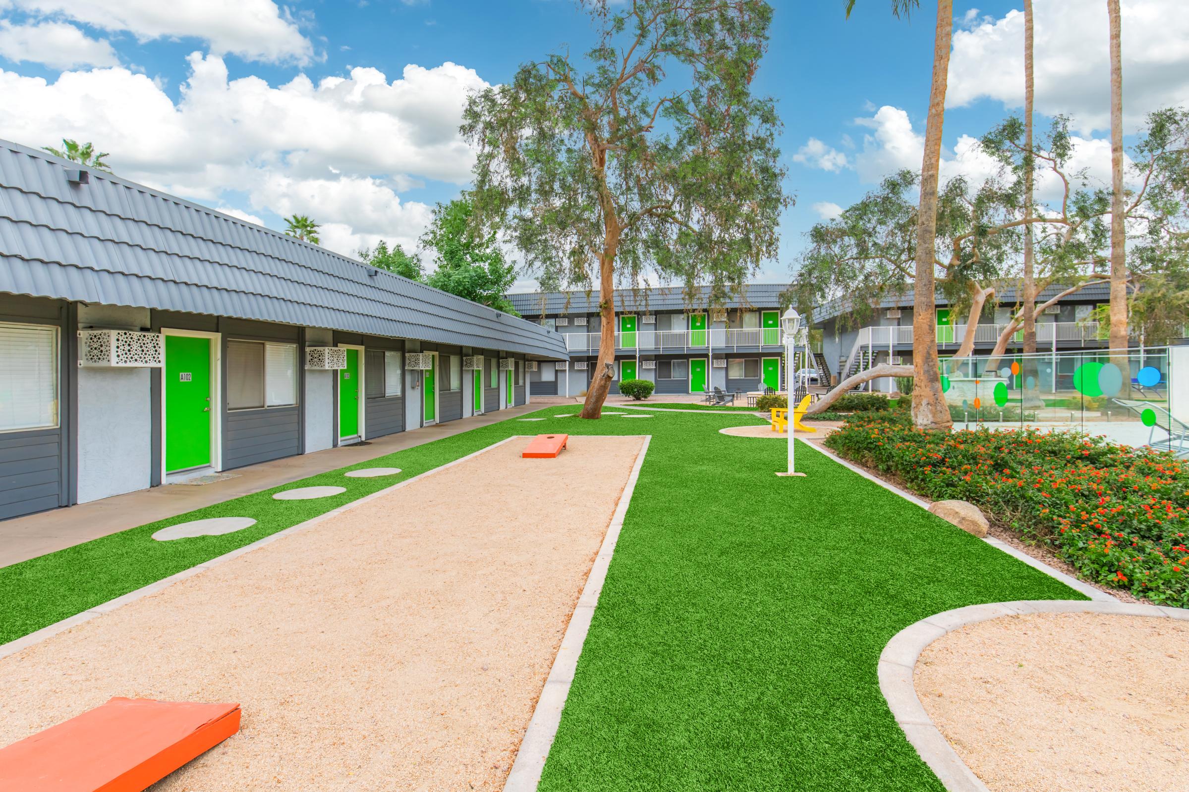 A vibrant courtyard of a modern hotel featuring green grass, sandy areas for games, colorful buildings with green accents, and palm trees under a blue sky with clouds. The setting is inviting and designed for relaxation and fun.
