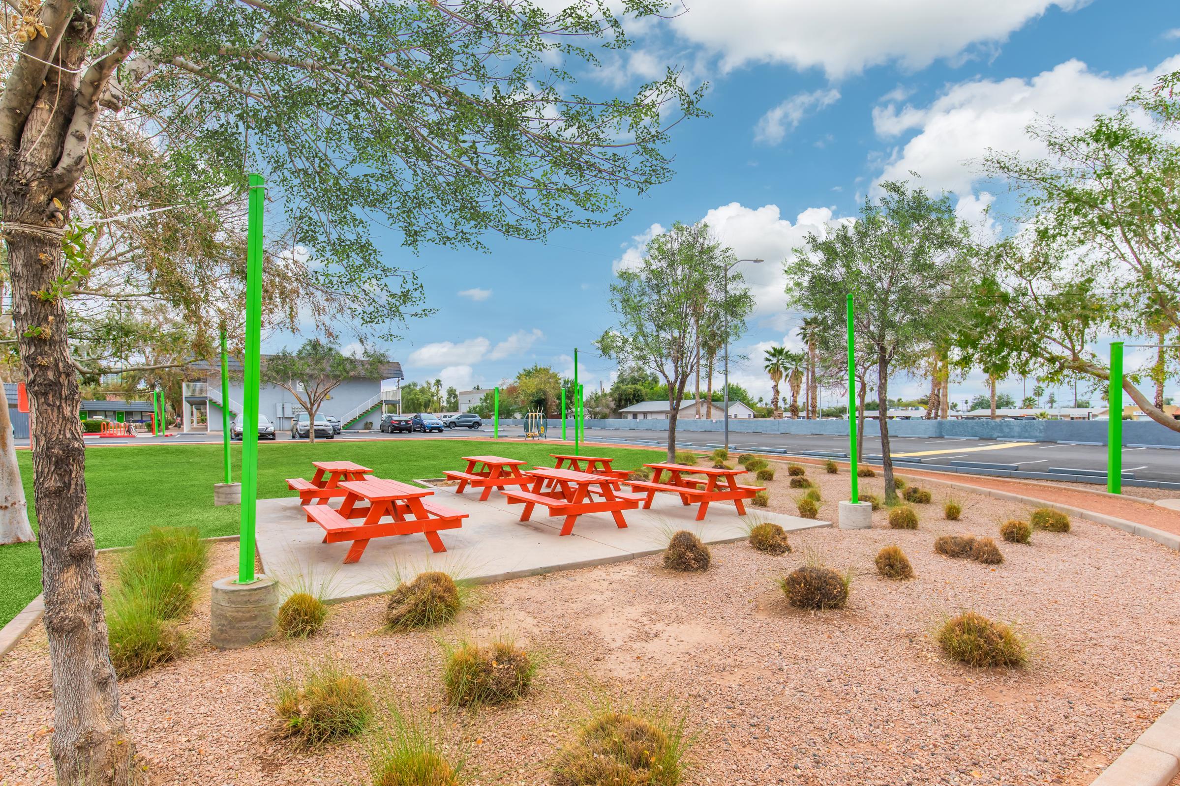 Colorful picnic tables in an outdoor area surrounded by greenery and gravel. Bright orange tables are arranged on a concrete pad, with green poles and sparse shrubbery. In the background, a parking lot and palm trees are visible under a partly cloudy sky.
