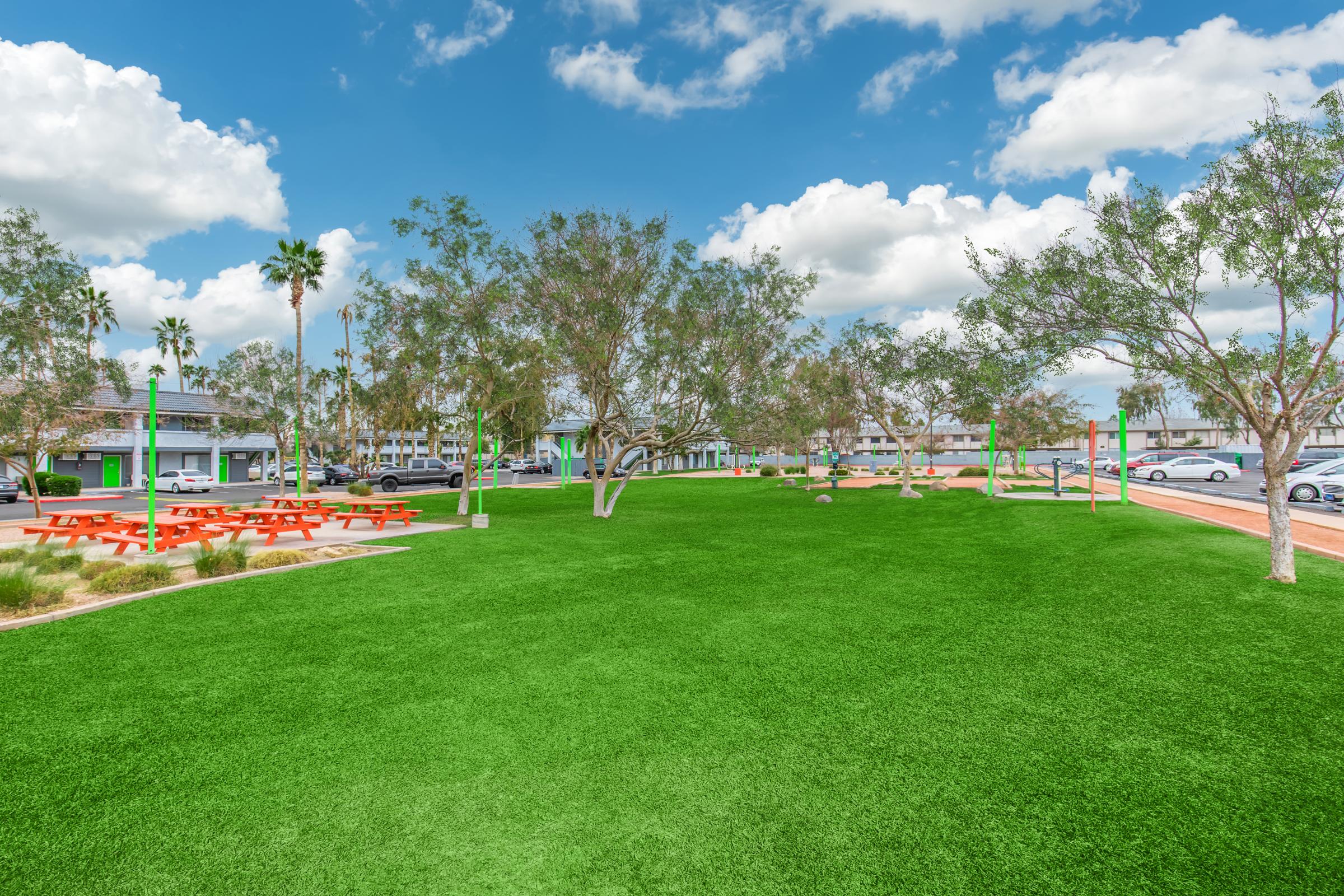 A bright, open park area featuring vibrant green artificial grass, with several small trees and picnic tables in the foreground. In the background, low-rise buildings and parked cars are visible under a partly cloudy blue sky. The scene conveys a spacious and welcoming outdoor environment.