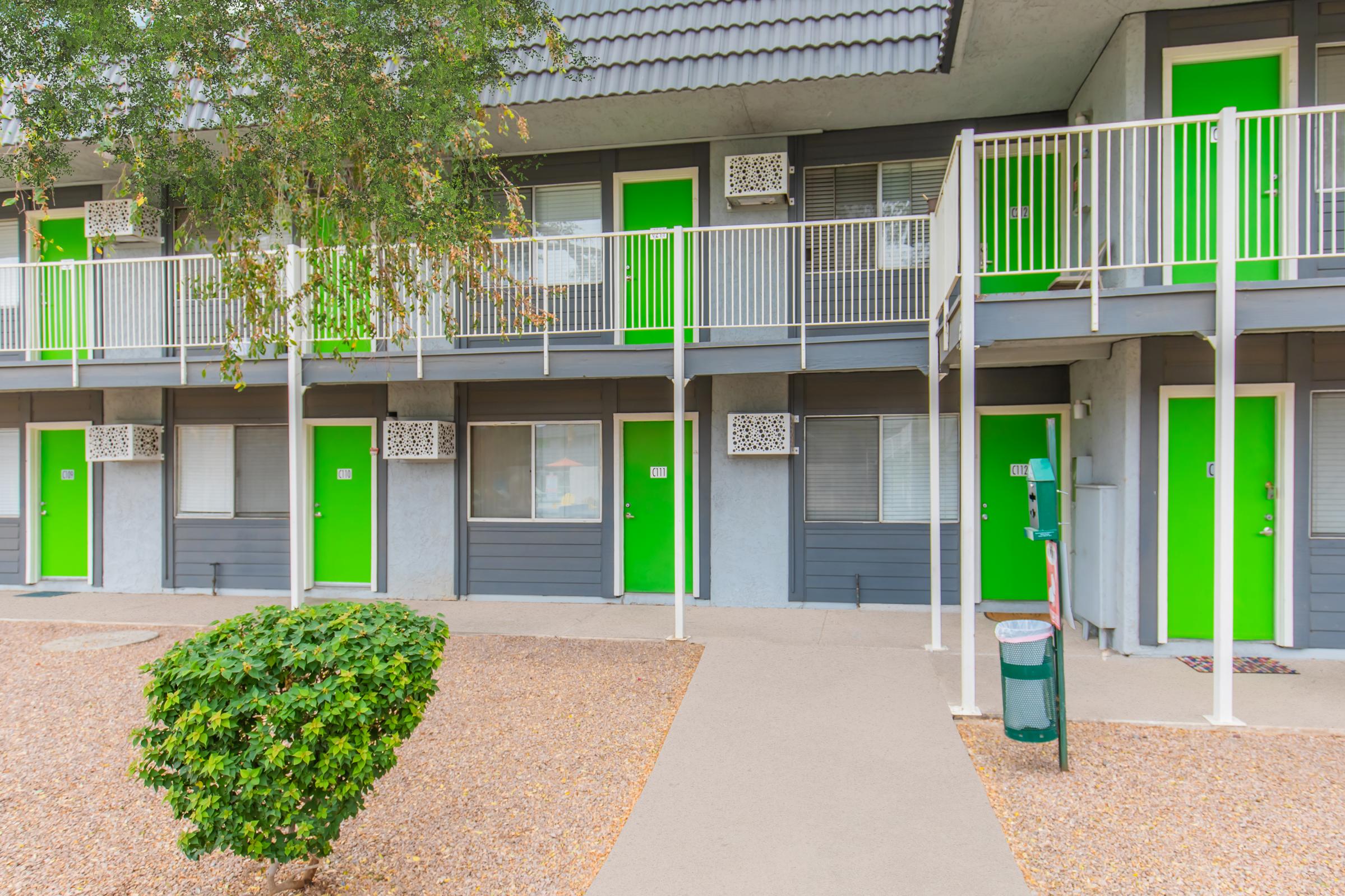 A row of motel-style rooms with vibrant green doors and grey siding. Each door has a small window above it, and a small shrub is visible in the foreground. There are metal railings on the second level balconies, creating a neat, organized appearance. The pathway is clear and leads to the doors.