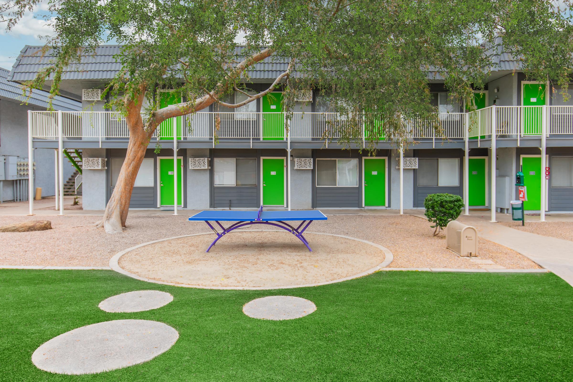 Colorful outdoor area featuring a ping pong table on grass, surrounded by gravel. In the background, multiple symmetrical buildings with green doors. A large tree provides some shade, creating a casual and inviting space for recreation.