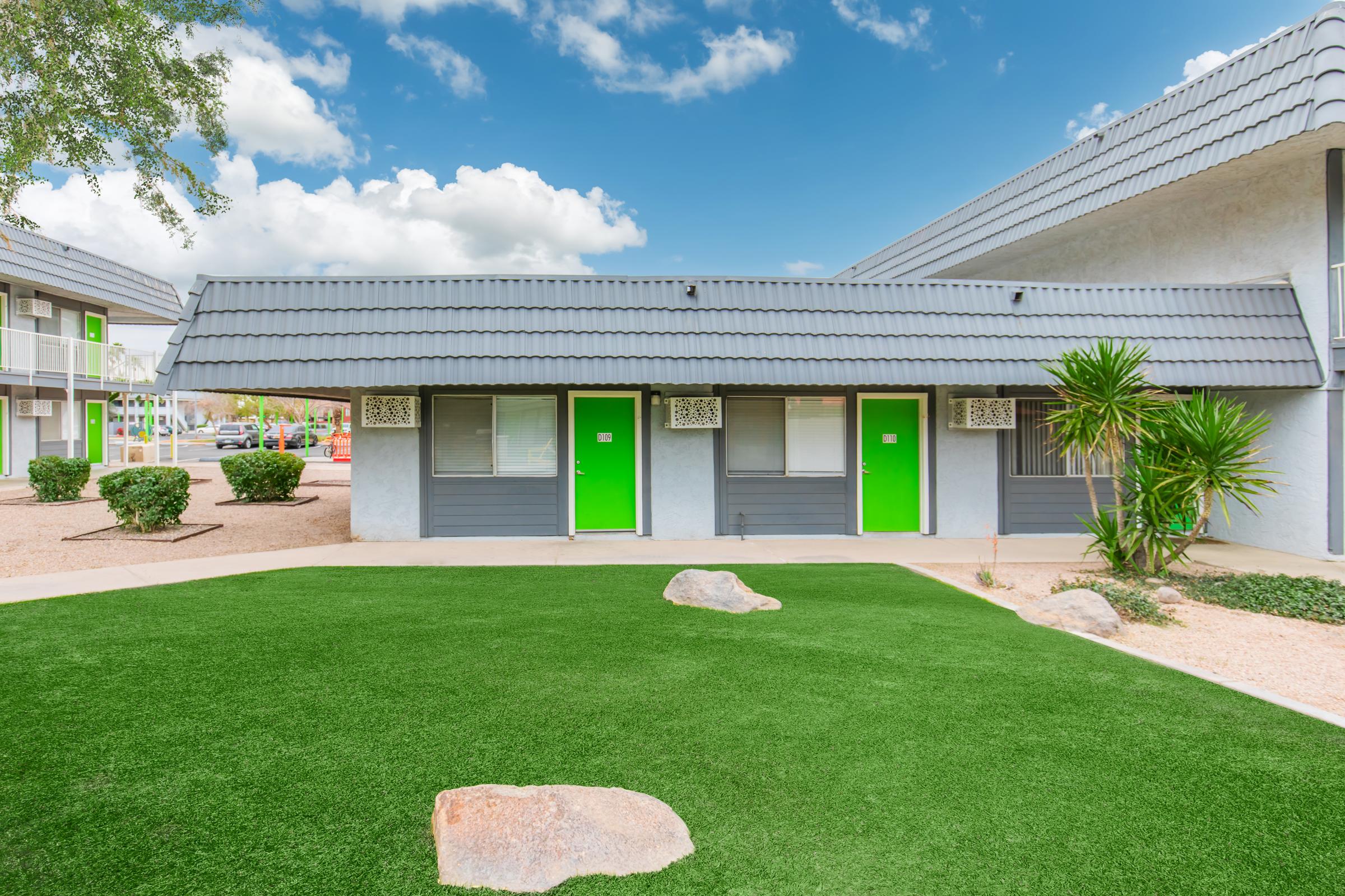 A low-rise building with gray exterior and triangular roof features several doors painted in vibrant green. Lush green artificial grass and a few rocks adorn the front area, with neatly trimmed shrubs in the background. The sky above is bright with scattered clouds, creating a pleasant outdoor atmosphere.