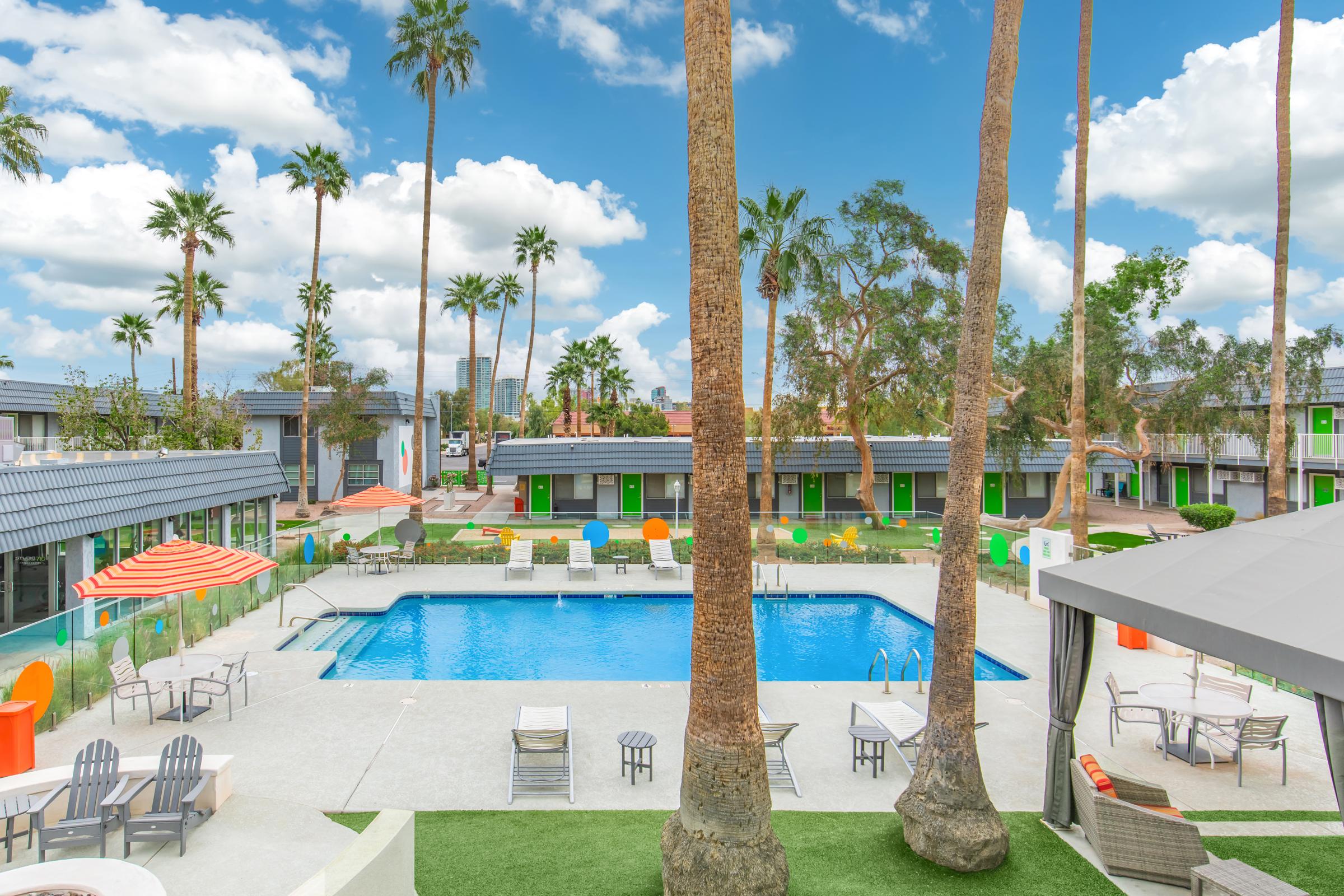 A vibrant pool area surrounded by palm trees, featuring lounge chairs and colorful seating. The pool is clear and inviting, with a shaded seating area nearby. Modern buildings with bright accents are visible in the background, set against a blue sky with fluffy clouds.