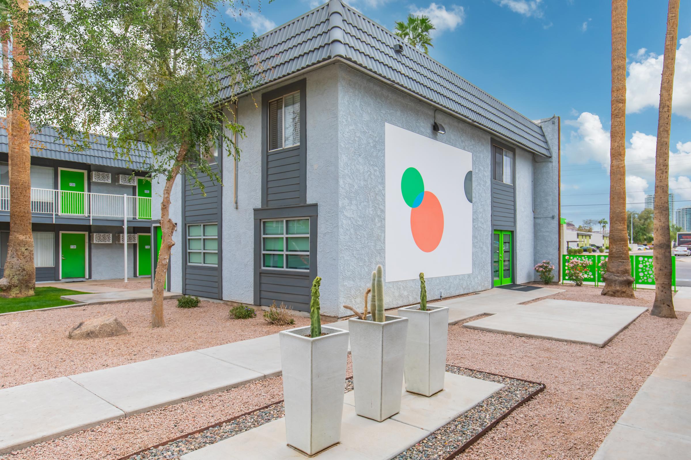 A modern building with a gray exterior and a large abstract mural featuring orange and green circles. The landscape includes palm trees and two planters with tall cacti. There are green doors and windows, and a clean pathway leading to the building, surrounded by decorative gravel.